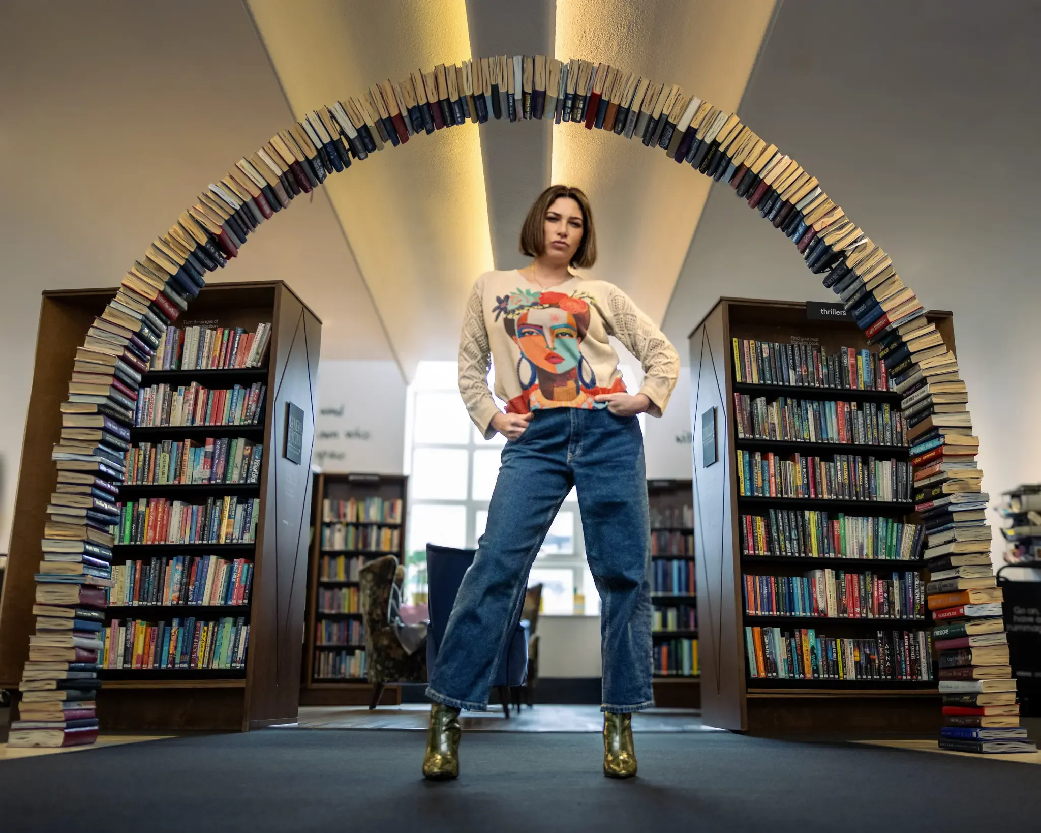 A woman stands confidently in a library, framed by an arch made of stacked books, with shelves of books behind her.
