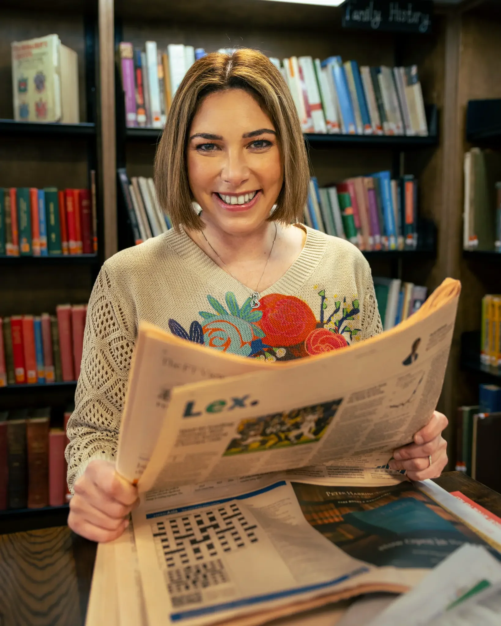 Young woman with shoulder-length hair smiling and holding a newspaper, sitting in a library with shelves of books in the background.