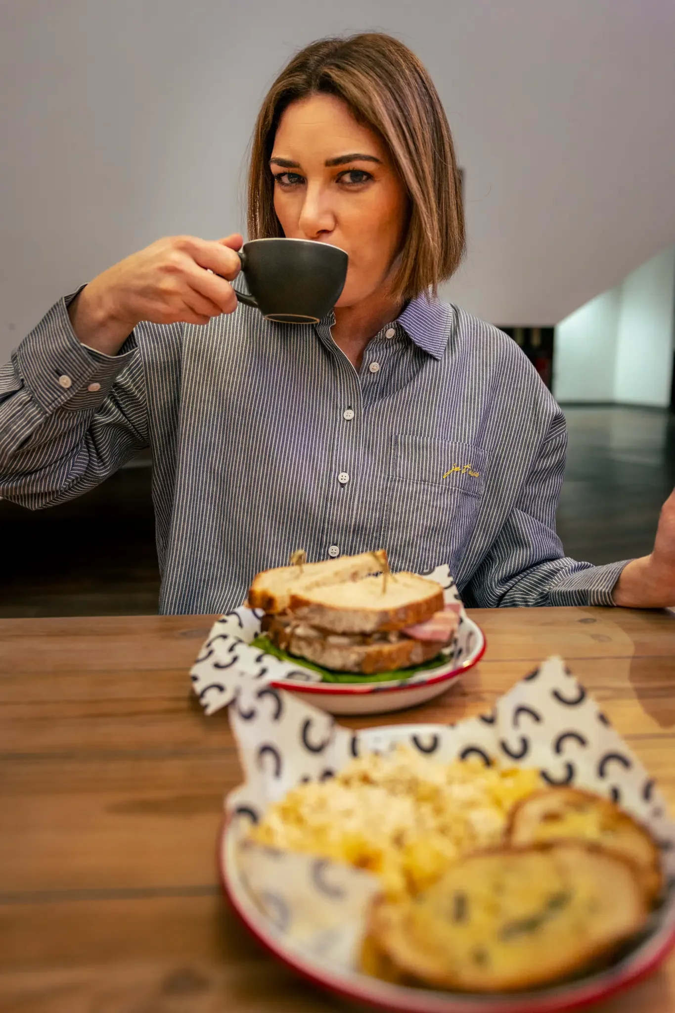 A woman with short brown hair in a striped shirt drinks coffee at a wooden table with a plate of sandwiches and a dish of baked dish or casserole.