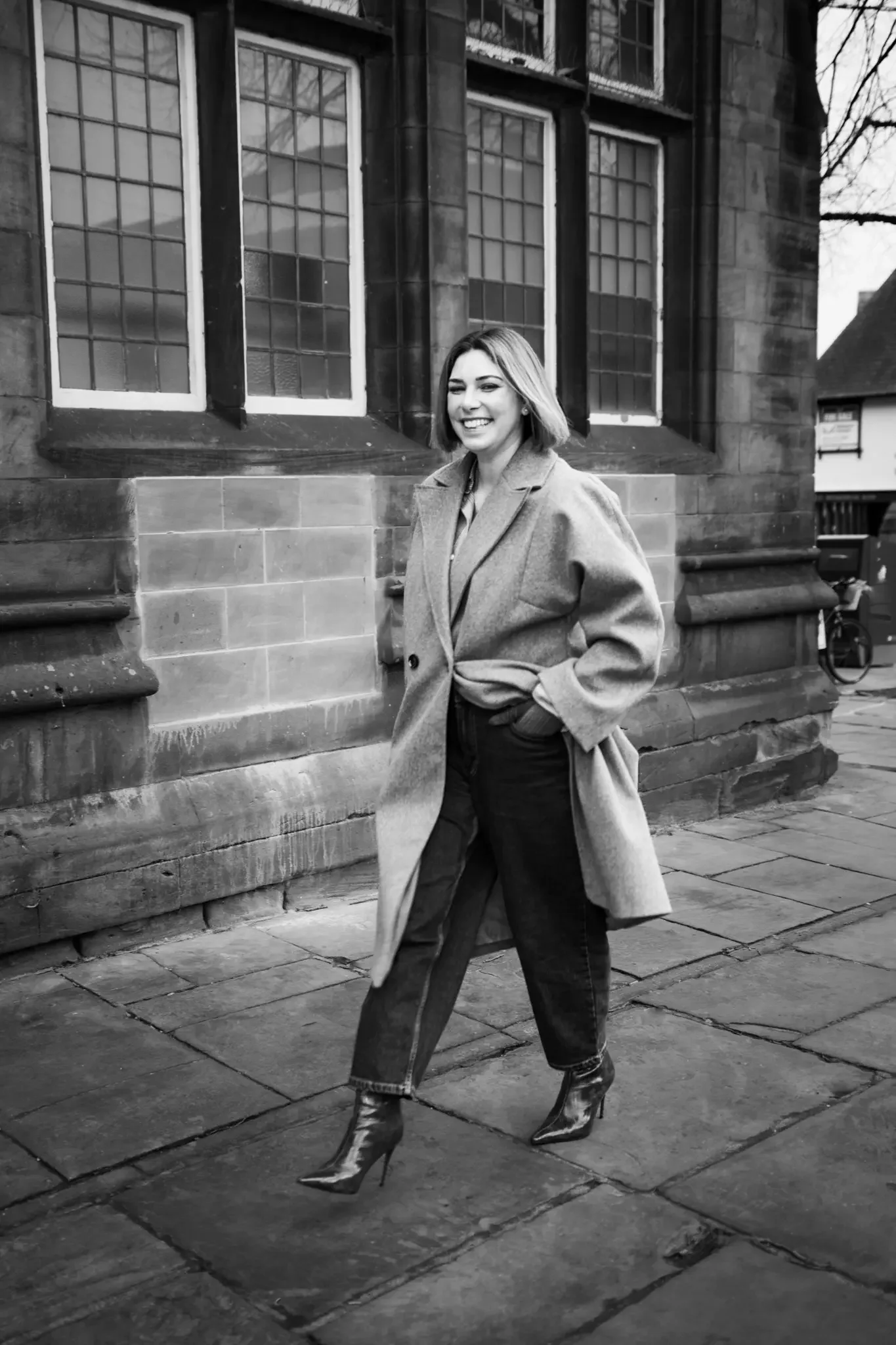 A woman walking outdoors past a historic building with large windows, smiling, wearing a coat, jeans, and high-heeled boots, in a black and white photo.