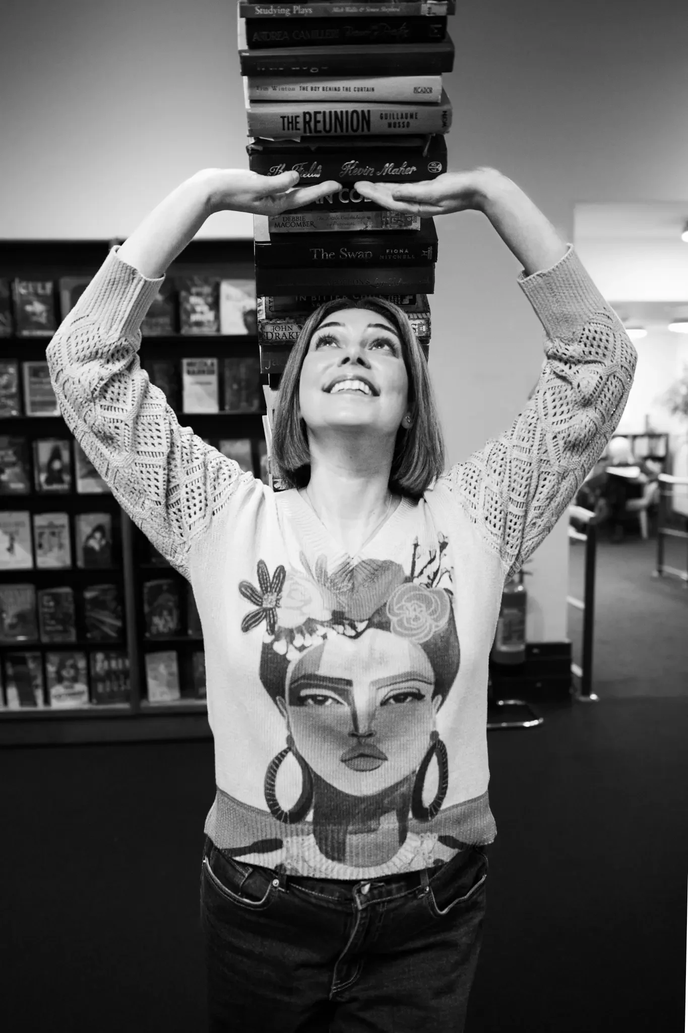 A woman holding a stack of books above her head in a bookstore. She is smiling and looking upward, wearing a Frida Kahlo sweater.