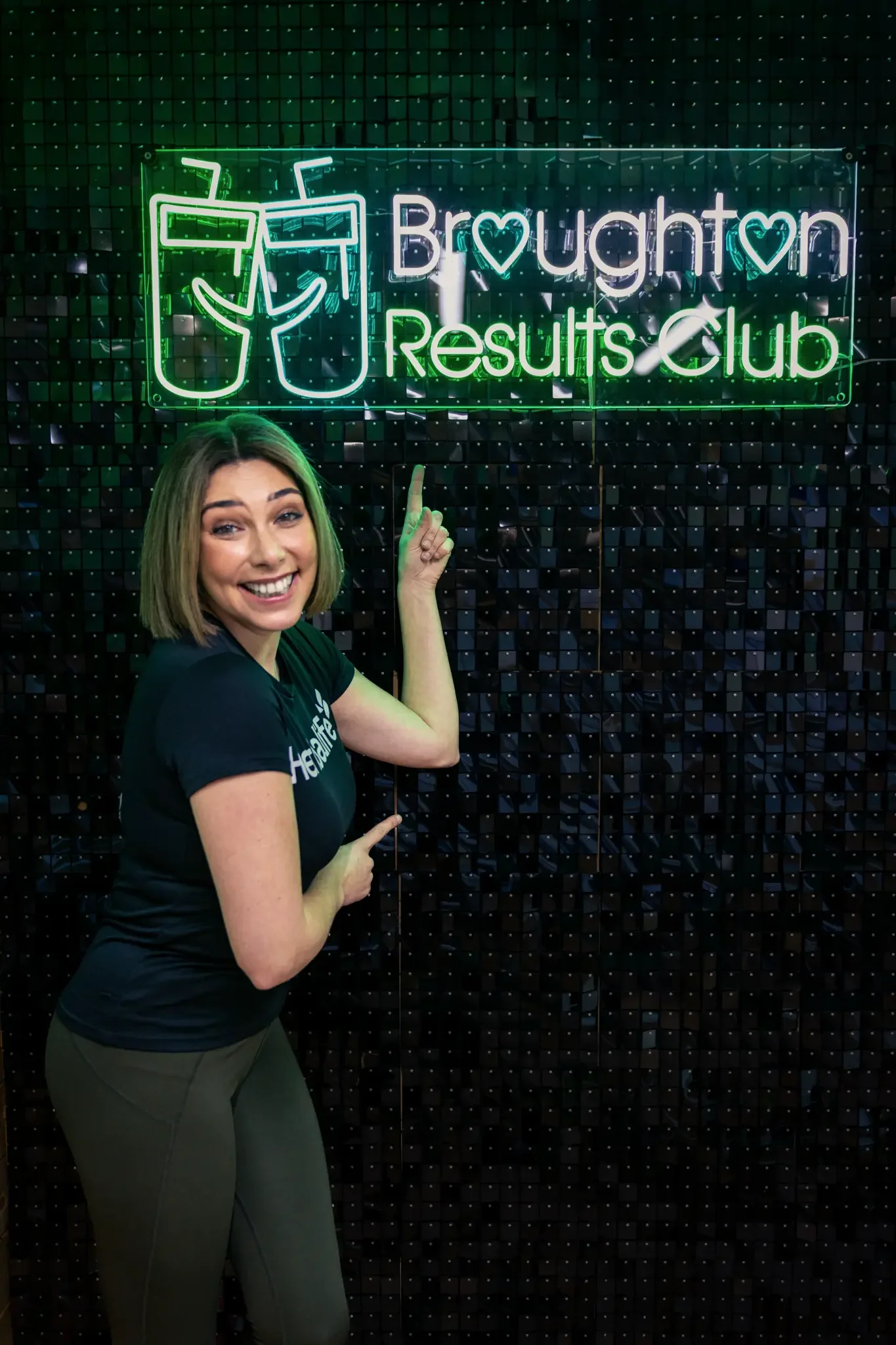 A smiling woman in a black T-shirt pointing to a neon sign that reads 'Broughton Results Club' with an image of two drinks, mounted on a dark, textured wall.