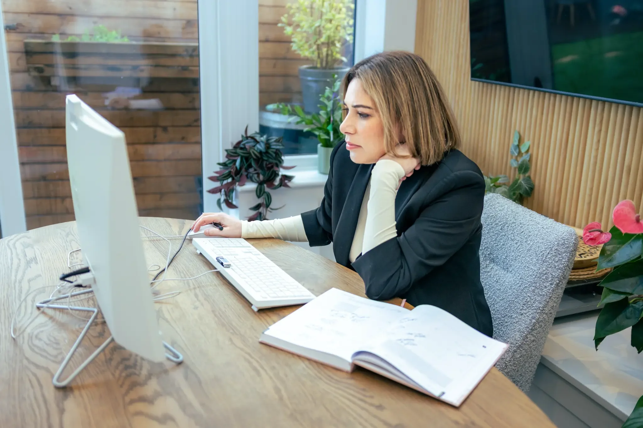 Woman sitting at a wooden desk working on a computer, with an open notebook nearby, plants in the background, and a large window.