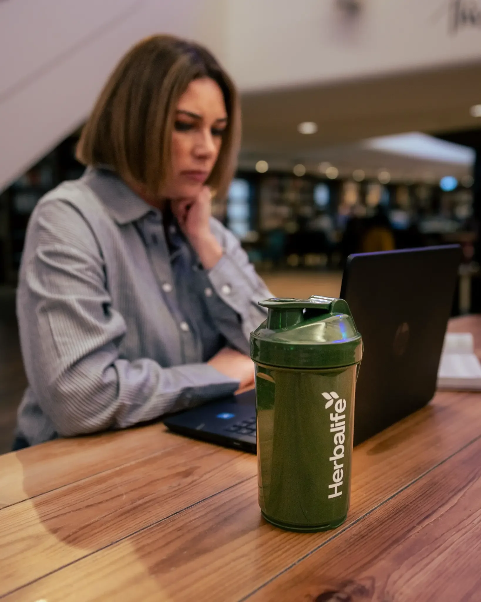 A woman sitting at a wooden table working on a laptop with a green HerbaLife shaker bottle in the foreground.