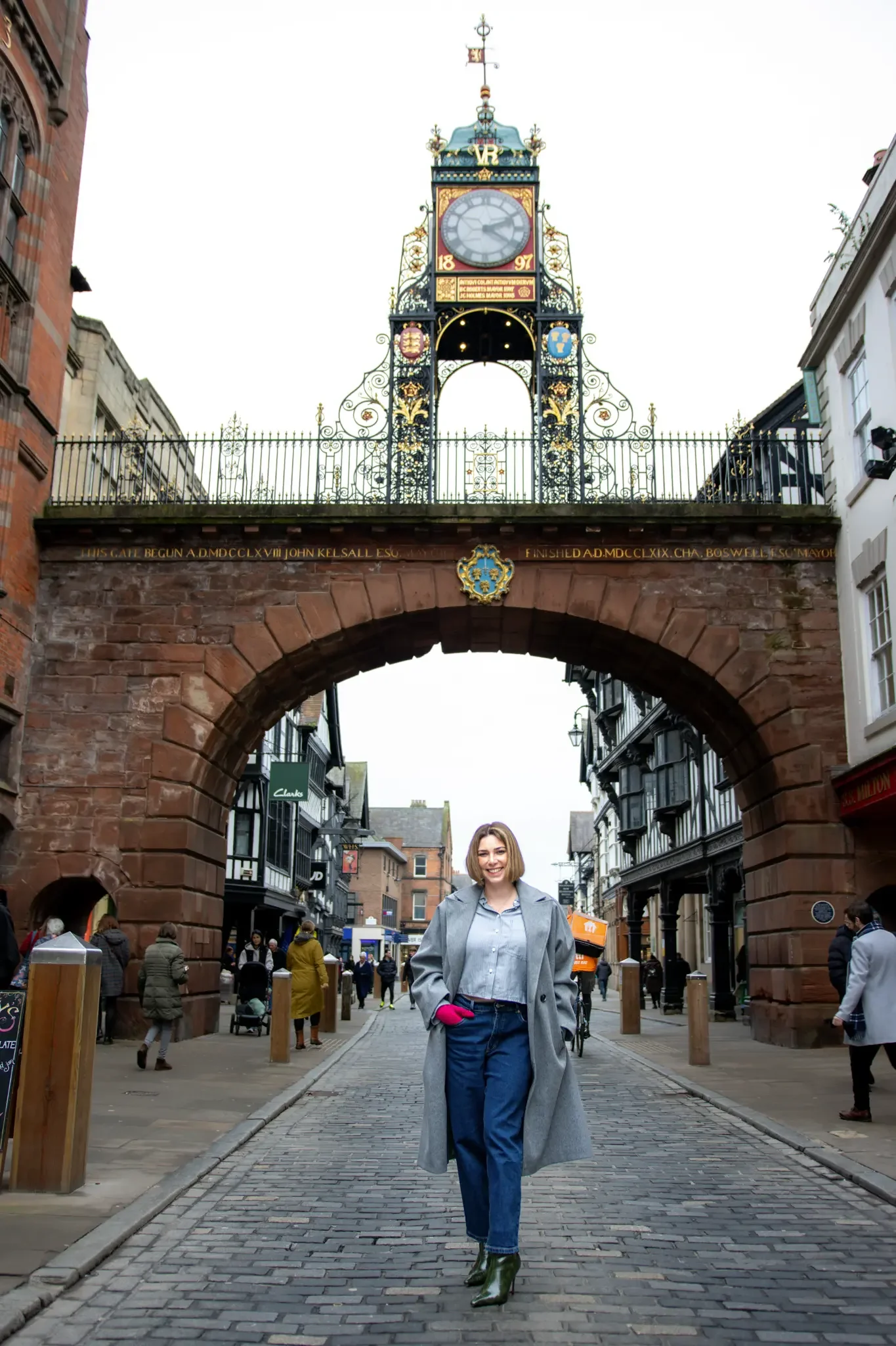 A woman standing on a cobblestone street in front of a historic archway with a clock tower above. The woman is smiling, wearing a gray coat, blue jeans, and green heeled boots.