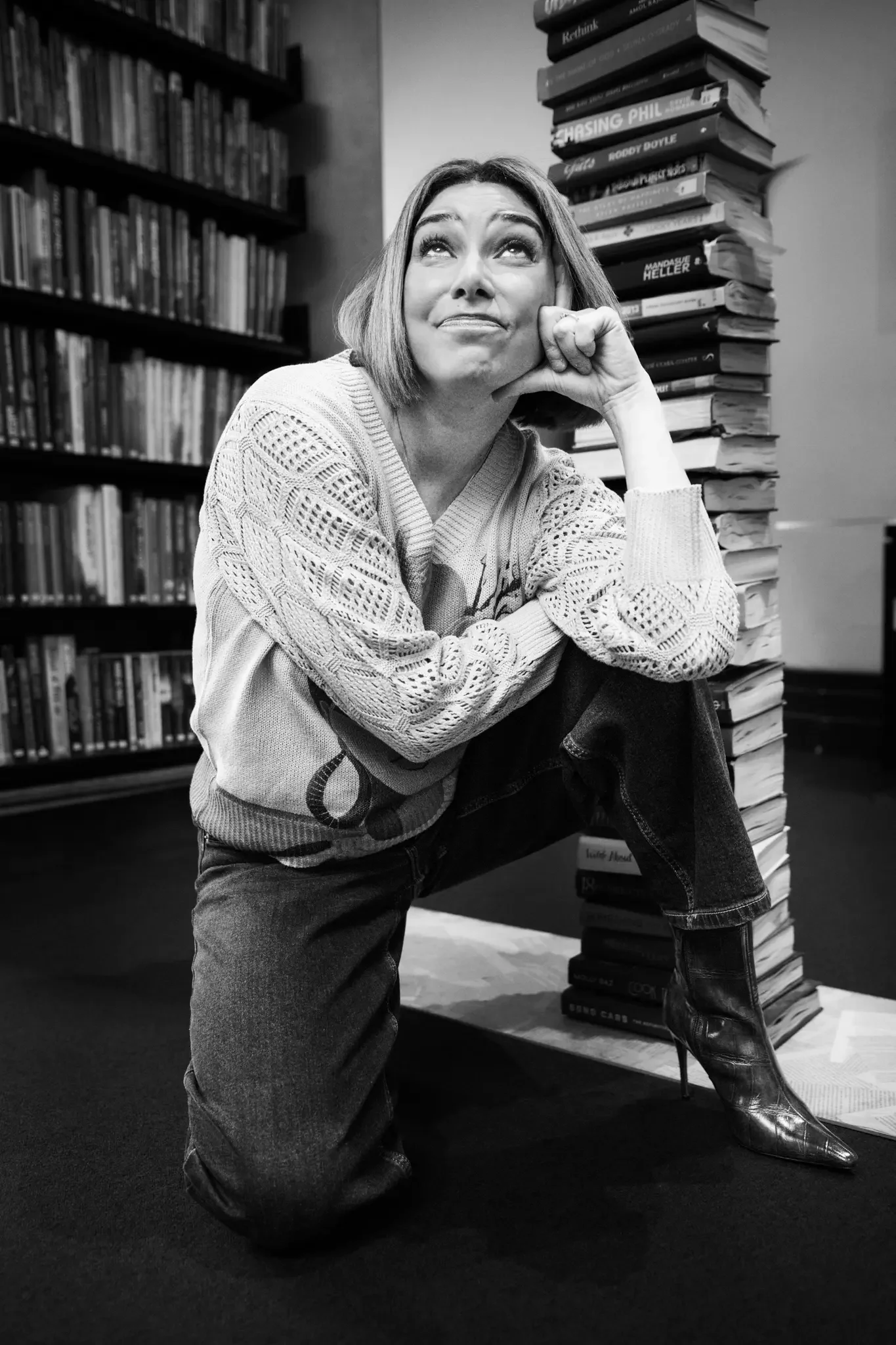 A woman with short hair and a patterned sweater kneels on the ground, resting her chin on her hand, in a room filled with bookshelves.