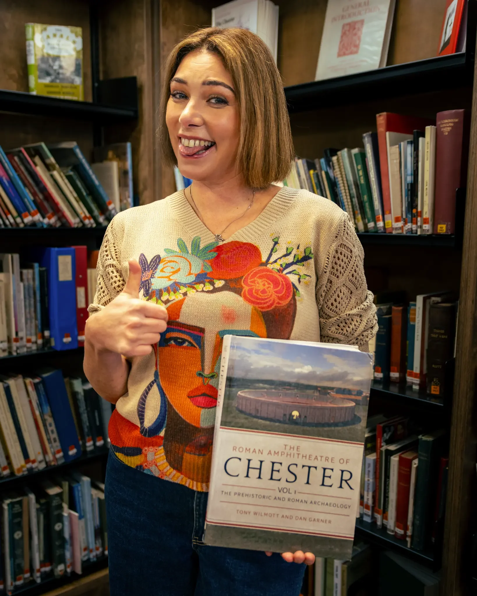 A woman standing in a library holding a book titled 'The Roman Amphitheatre of Chester', giving a thumbs-up and sticking her tongue out playfully.