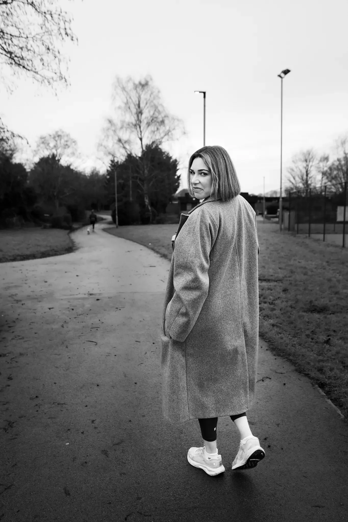 A woman in a coat walking on a park path, looking back over her shoulder, with trees and a person in the distance.