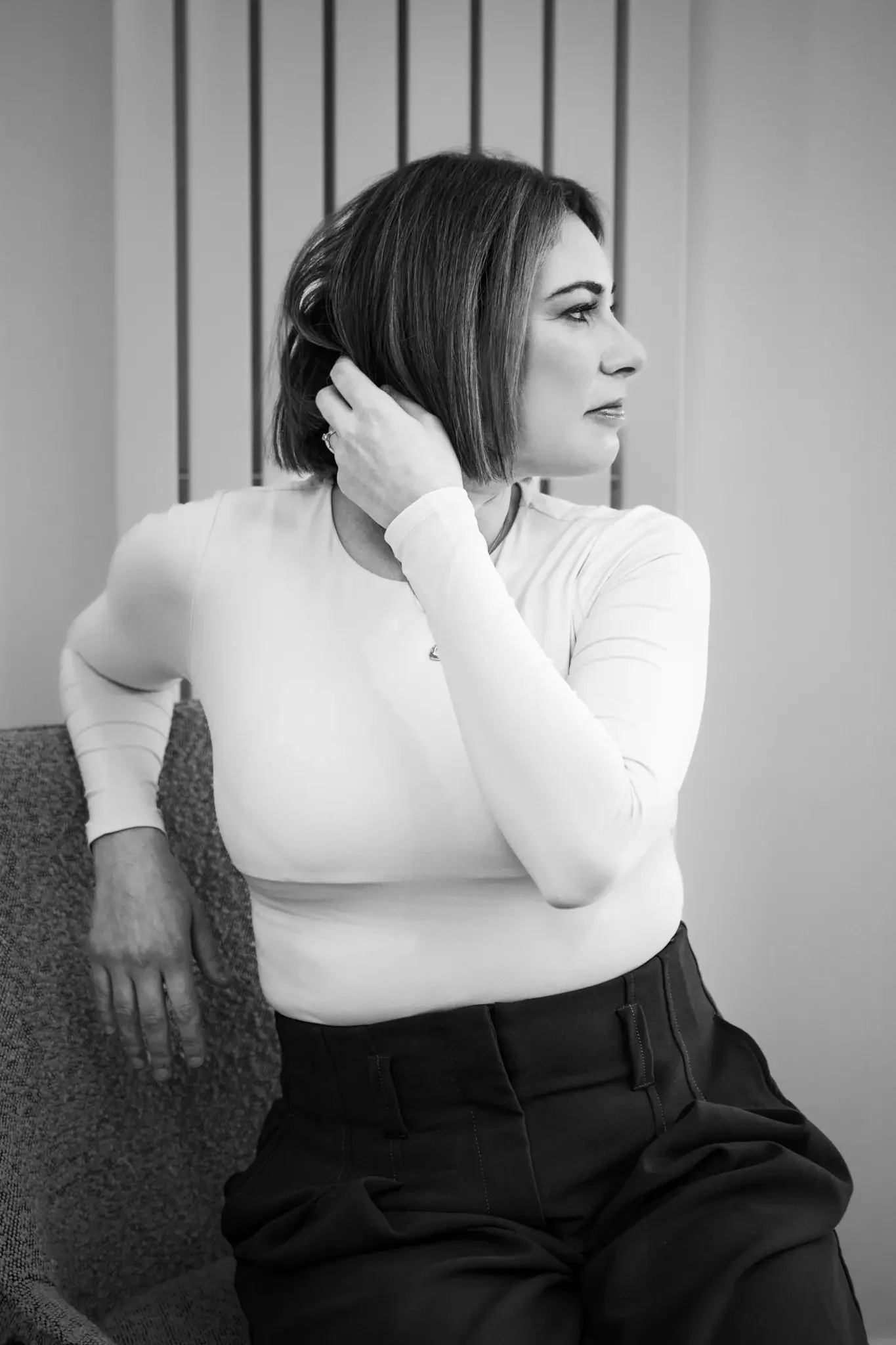 A woman in a white long-sleeve shirt and black pants sitting on a textured chair, touching her short dark hair with her right hand, looking to the right in a room with vertical wall panels.