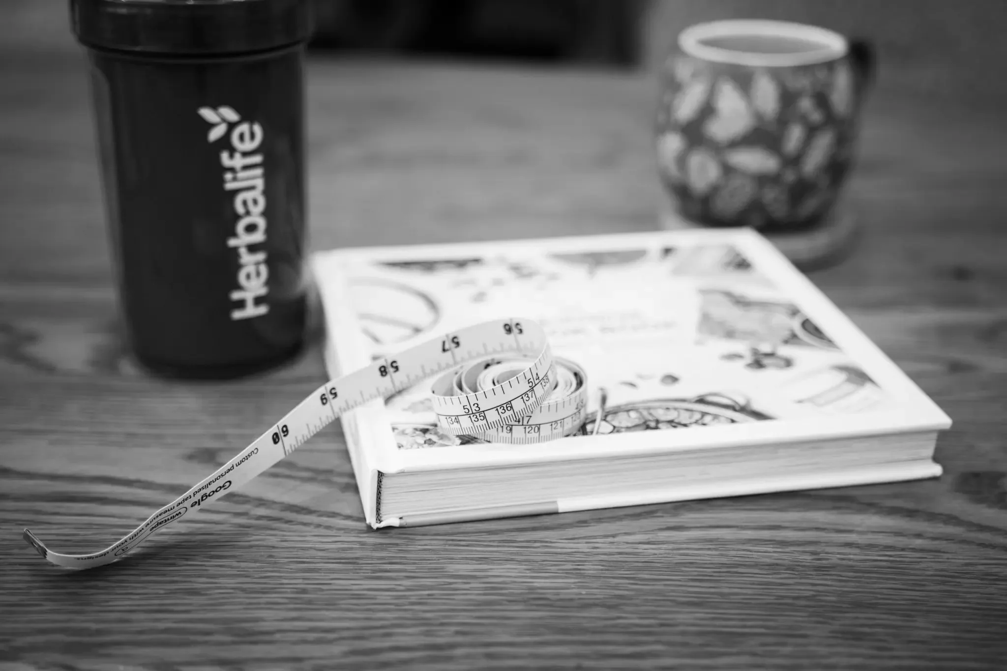 A black reusable coffee tumbler with the 'Herbalife' logo, a packet of colorful candies, a comic book, a measuring tape, and a patterned cup on a wooden table.