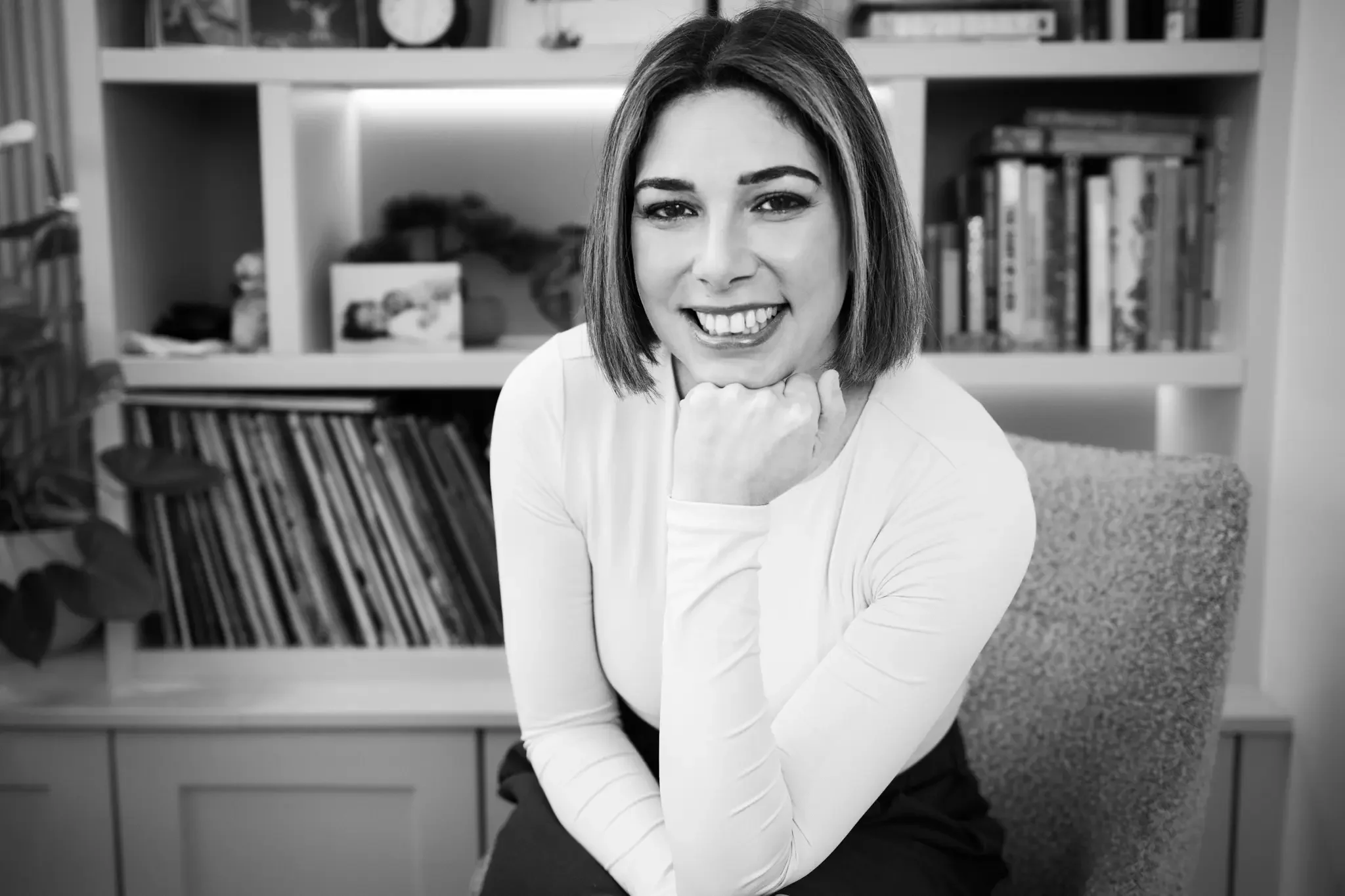 A smiling woman with short dark hair, wearing a white long-sleeve shirt, sitting in front of a bookshelf filled with books and decorative items, resting her chin on her right hand.