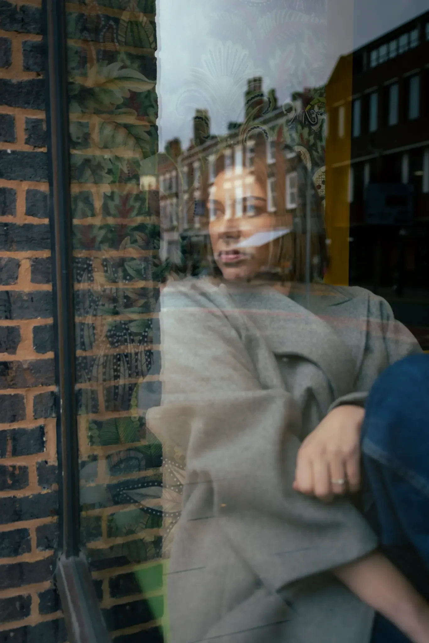 A woman sitting by a window with a reflection of city buildings outside. She has shoulder-length hair and is wearing a gray sweater, gazing outside contemplatively.