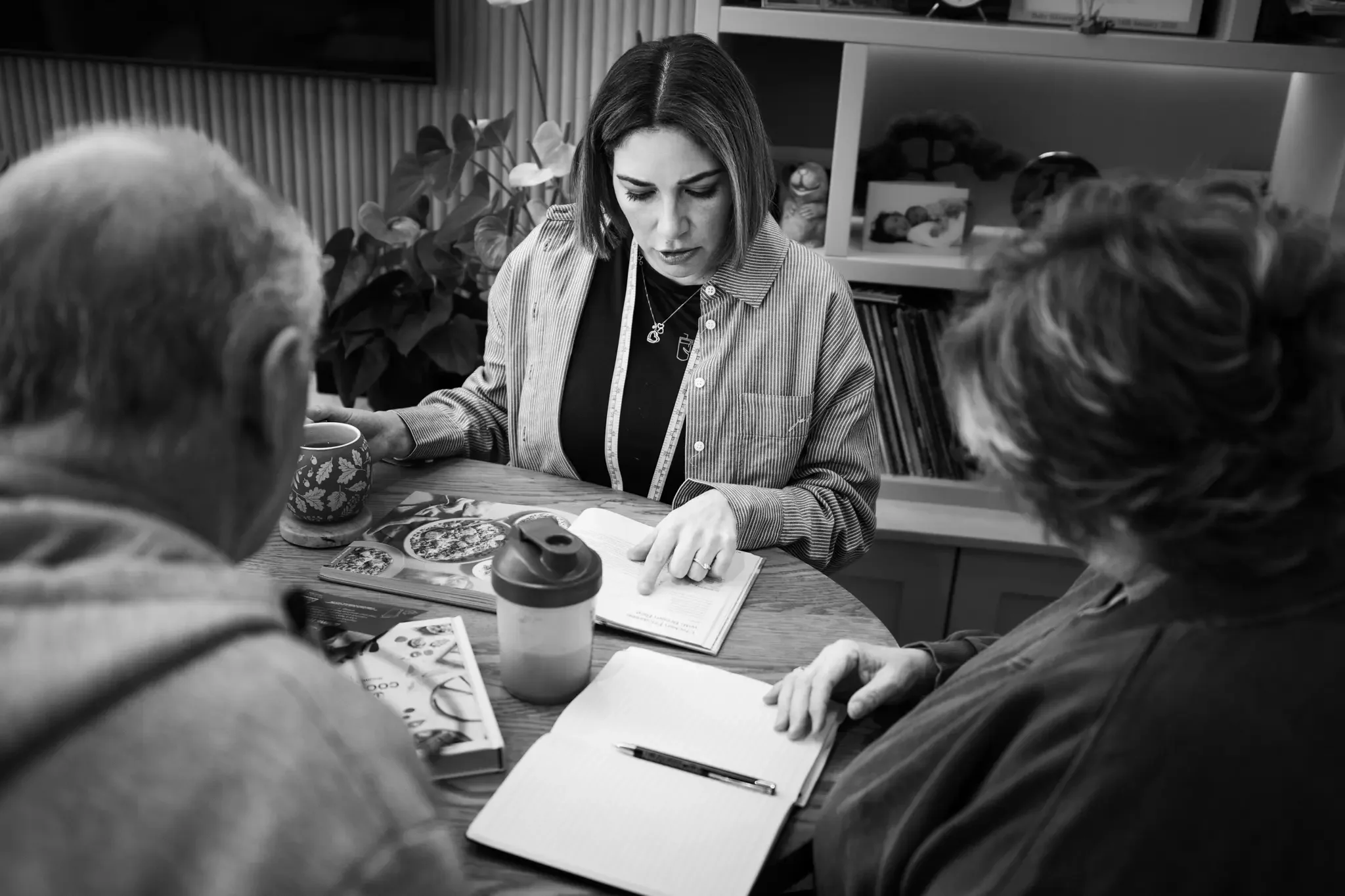 A woman explaining something to two elderly people at a table, with notebooks and magazines, in a room with shelves of books and decorative items.