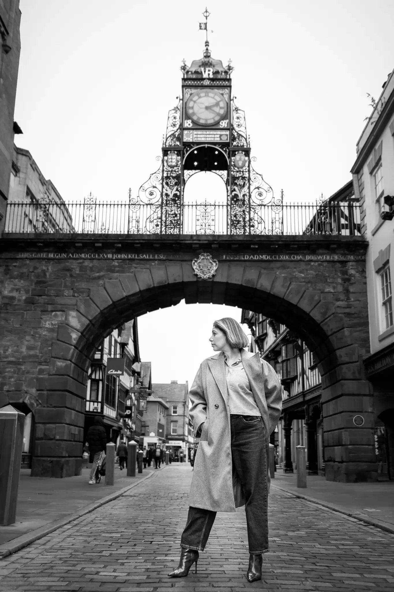 A woman standing on a cobblestone street beneath an ornate clock tower, with buildings lining the street, in black and white.