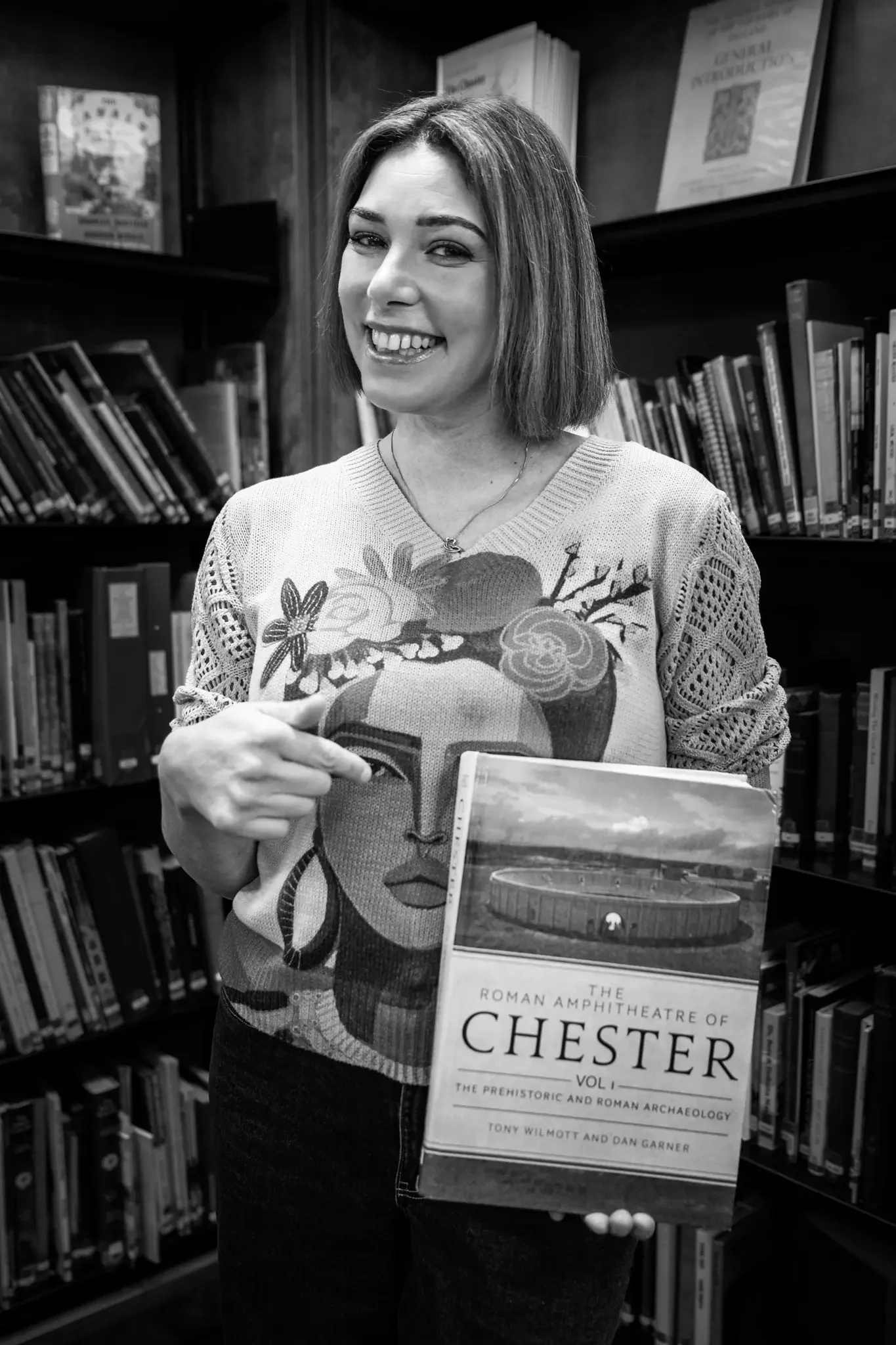 A smiling woman with short hair pointing to a book titled 'The Roman Amphitheatre of Chester Vol. 1: The Prehistoric and Roman Archaeology' in a library with shelves of books in the background.