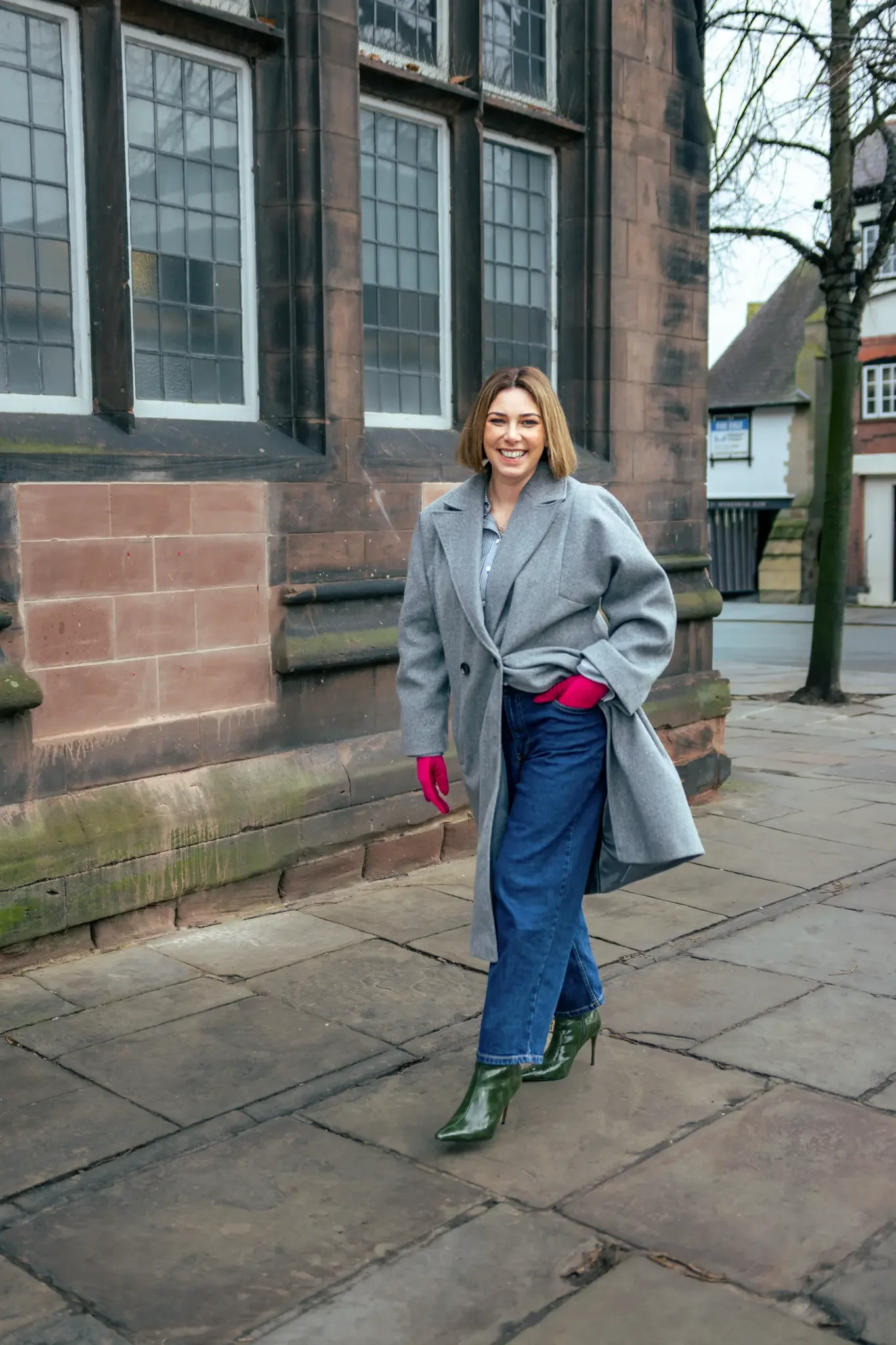 A woman with short brown hair wearing a gray coat, blue jeans, green high-heeled boots, and pink gloves stands outside on a stone sidewalk near a historic brick building with large window panes. She is smiling at the camera.