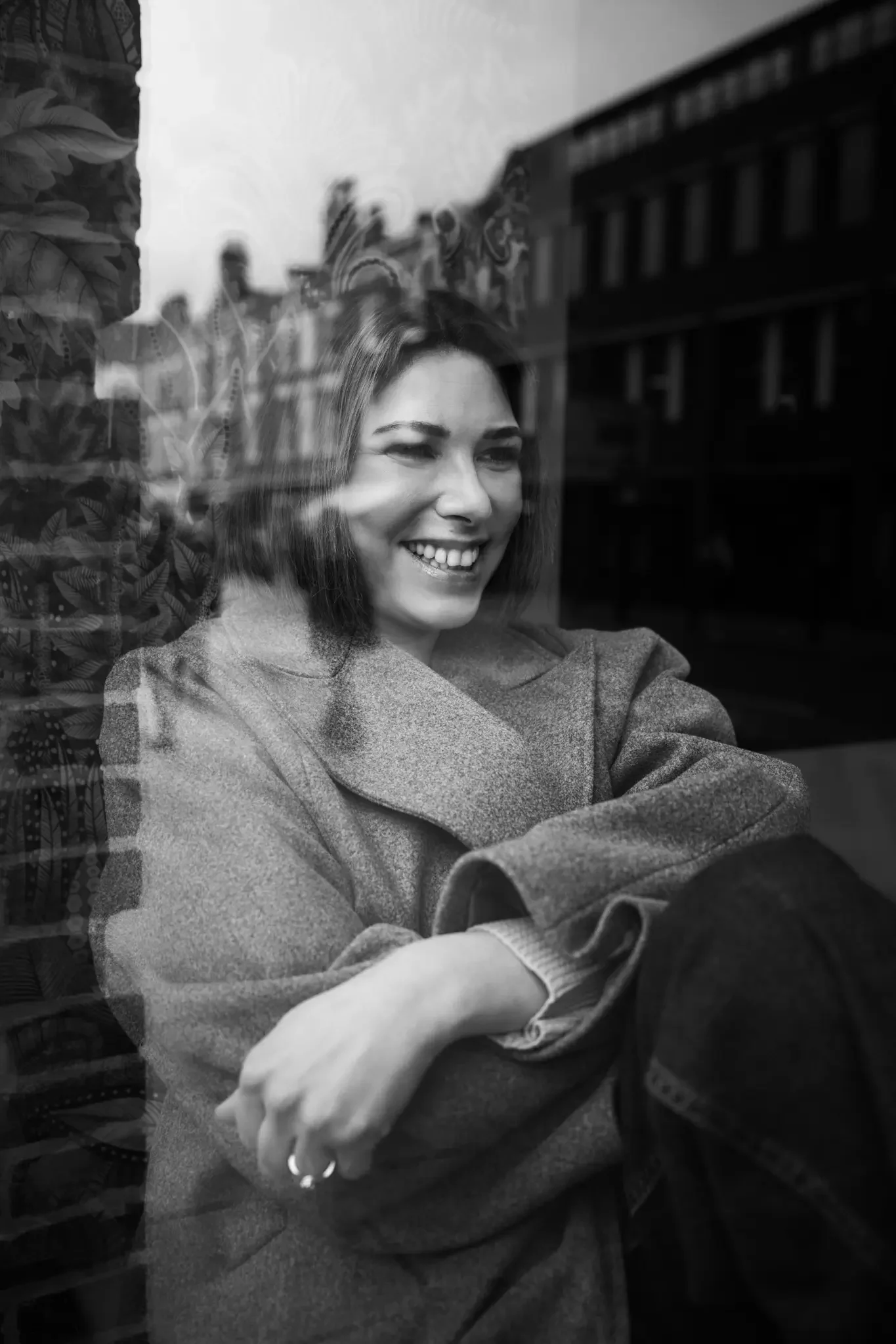 A woman with short hair and a coat sitting by a window, smiling as she looks outside. The photo is in black and white, reflecting buildings and trees outside.
