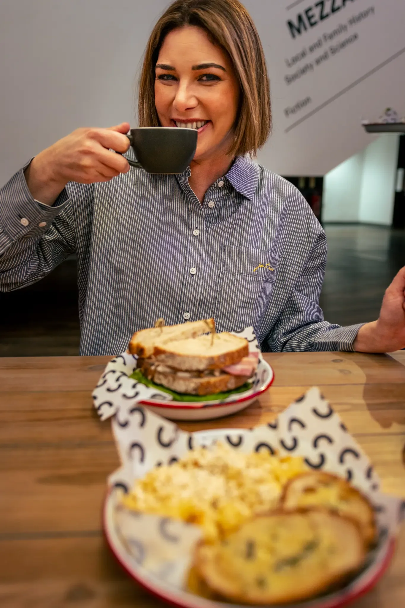 Woman with short brown hair enjoying a cup of coffee or tea at a restaurant table with a sandwich and a plate of egg dishes in front of her.