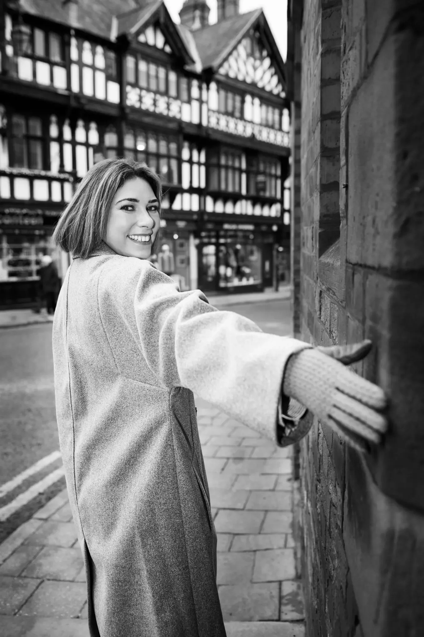 A woman in a coat and gloves smiling and reaching out her hand toward a brick wall on a city street with buildings in the background.