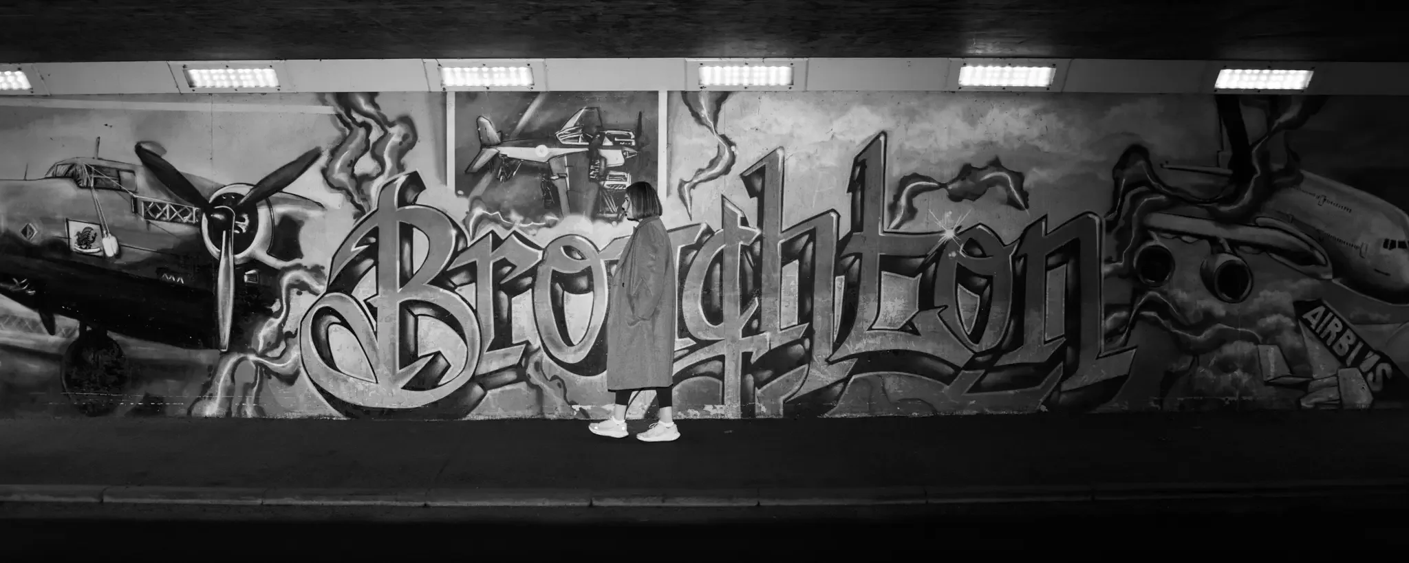 A person in a coat and sneakers standing in front of a graffiti mural with airplanes and the word 'Brodington' in stylized lettering.