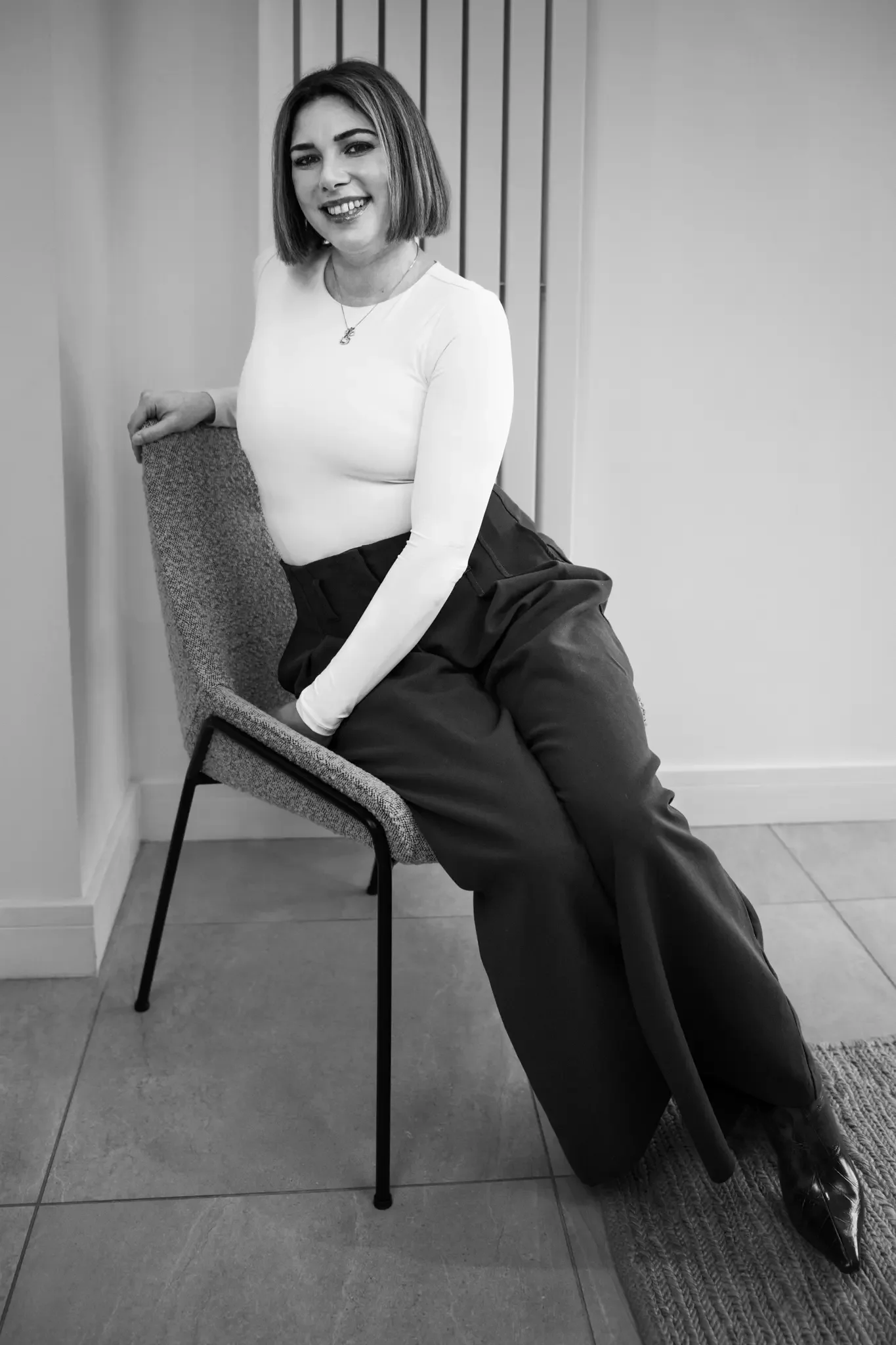 A smiling woman with shoulder-length hair sitting on a chair, leaning against the backrest, in a room with tiled floor and a plain wall.