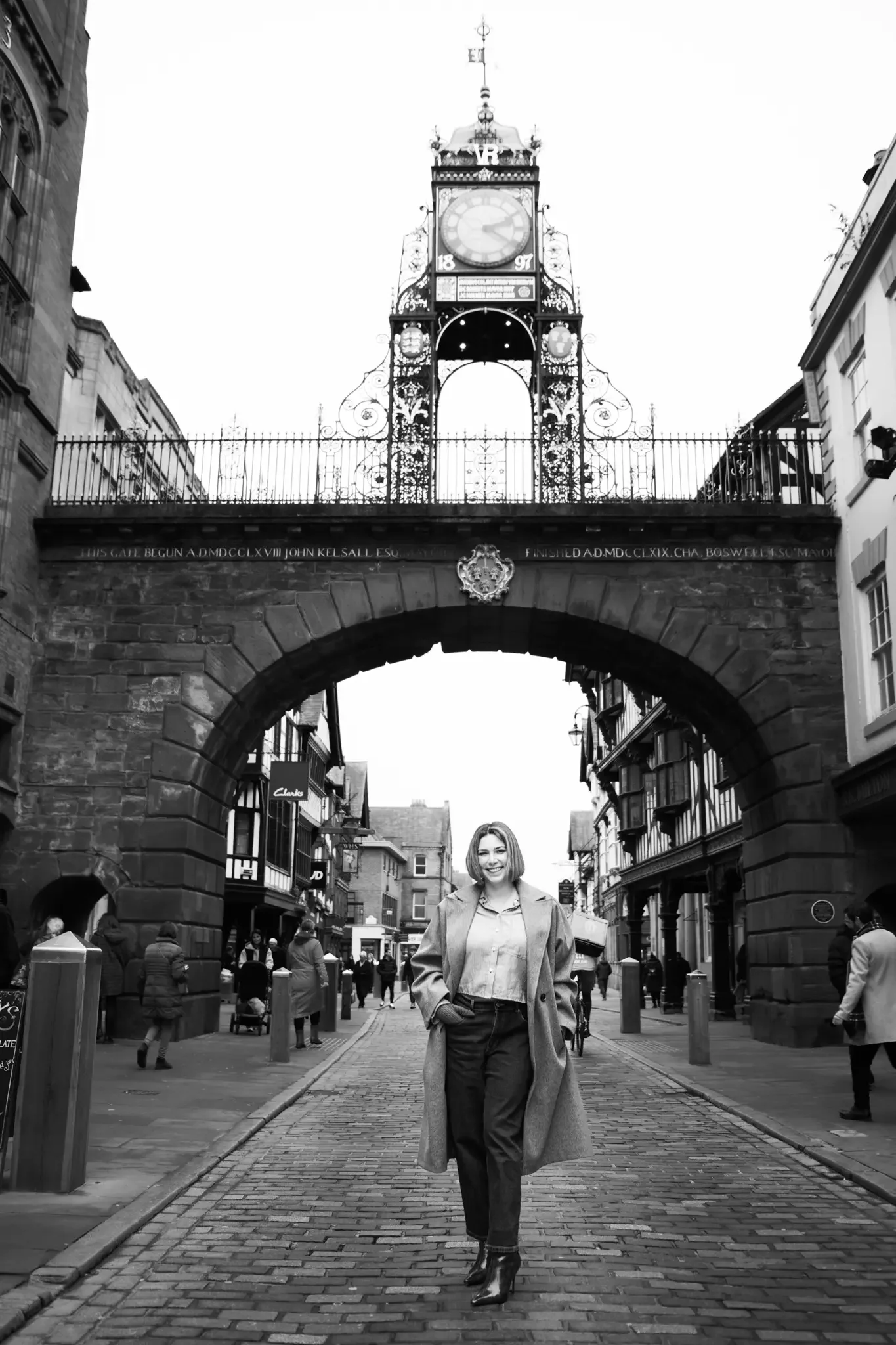A woman walking on a cobblestone street under a decorative arched bridge with an ornate clock tower above, in an urban setting.