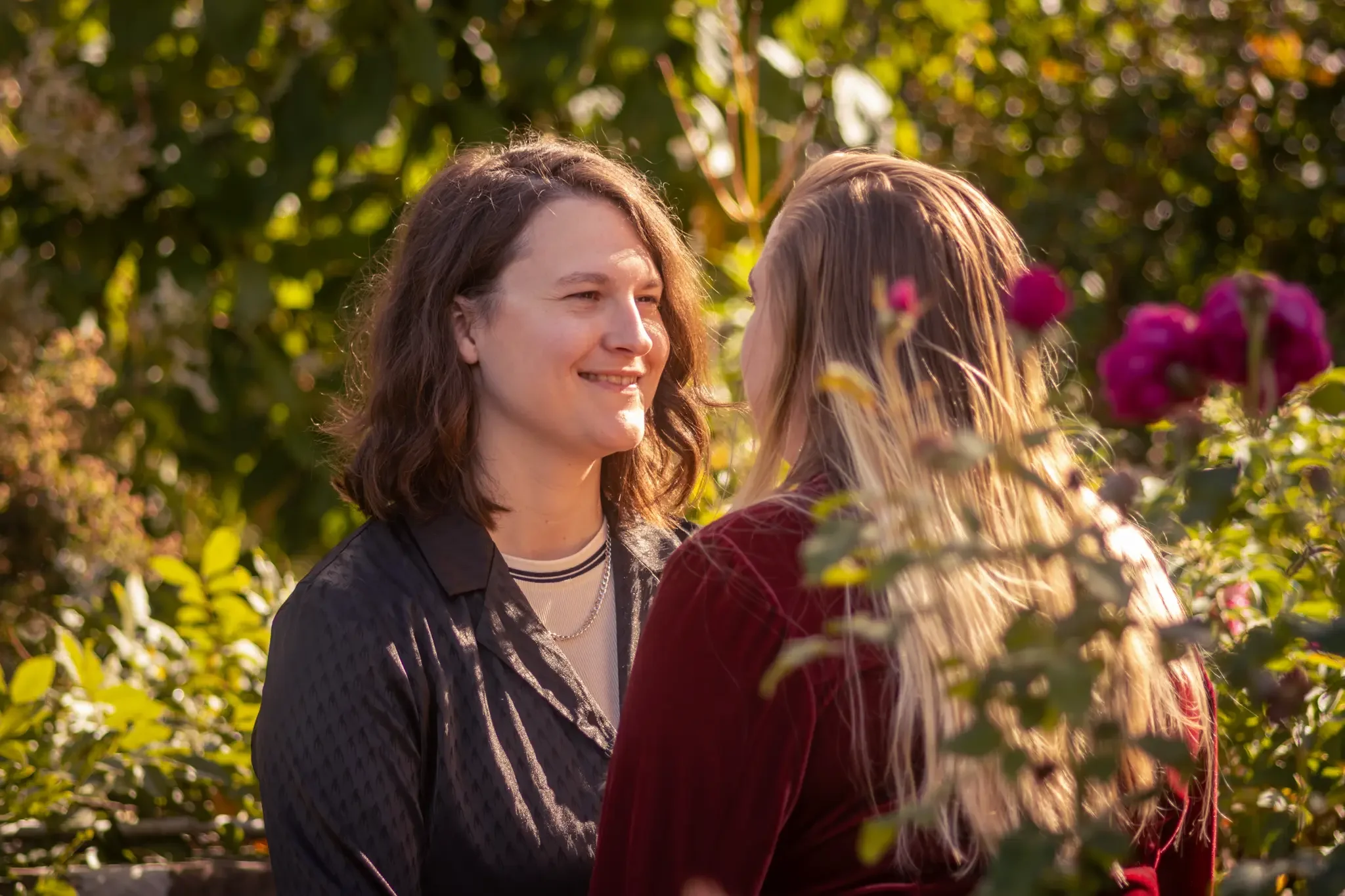 Two women standing outdoors in a garden or park, smiling and facing each other, with greenery and flowers around them, during golden hour.
