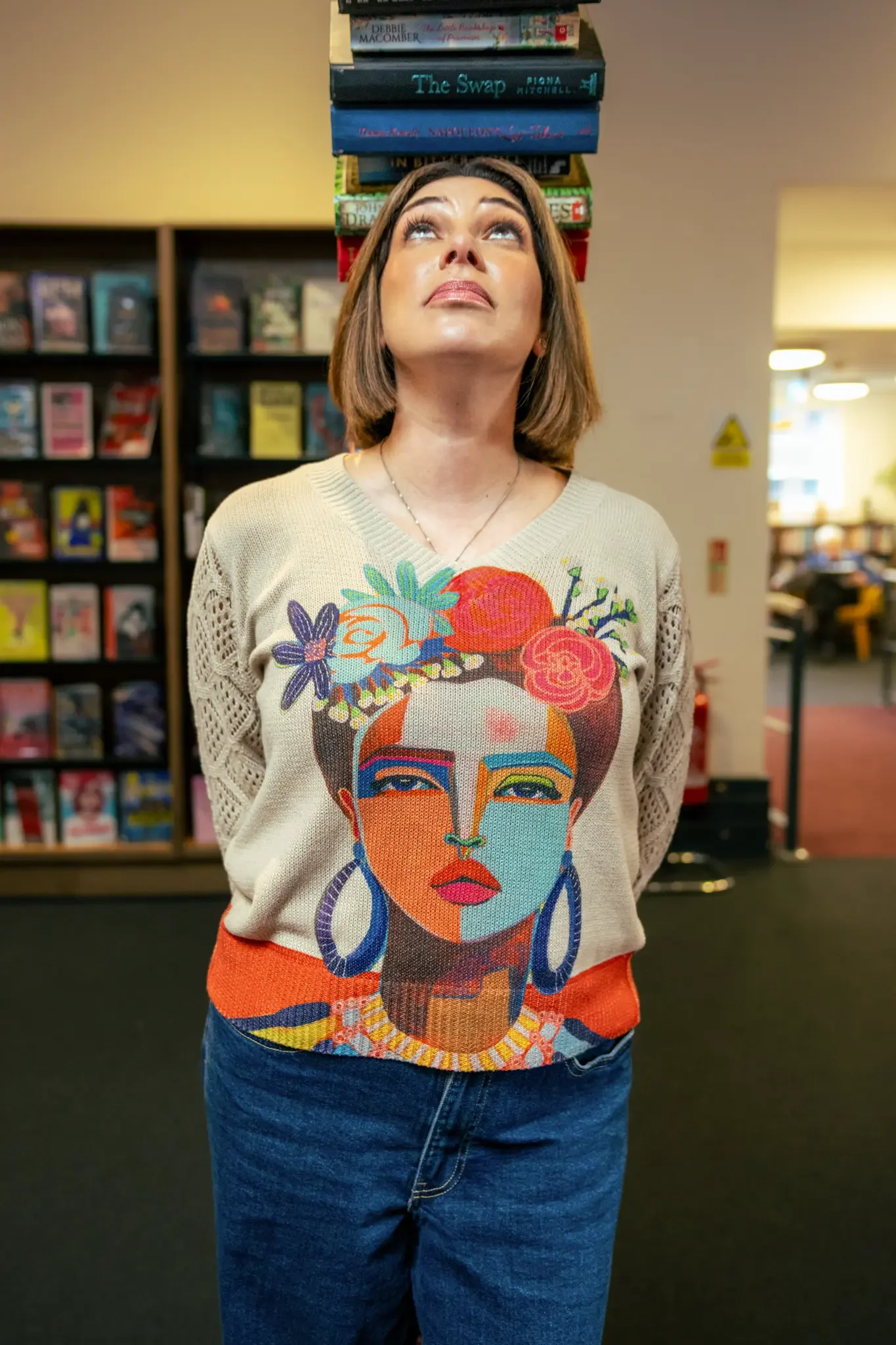 A woman with short brown hair looking up in a bookstore, balancing a stack of books on her head. She wears a beige sweater with a colorful, artistic portrait of a woman adorned with flowers and jewelry.