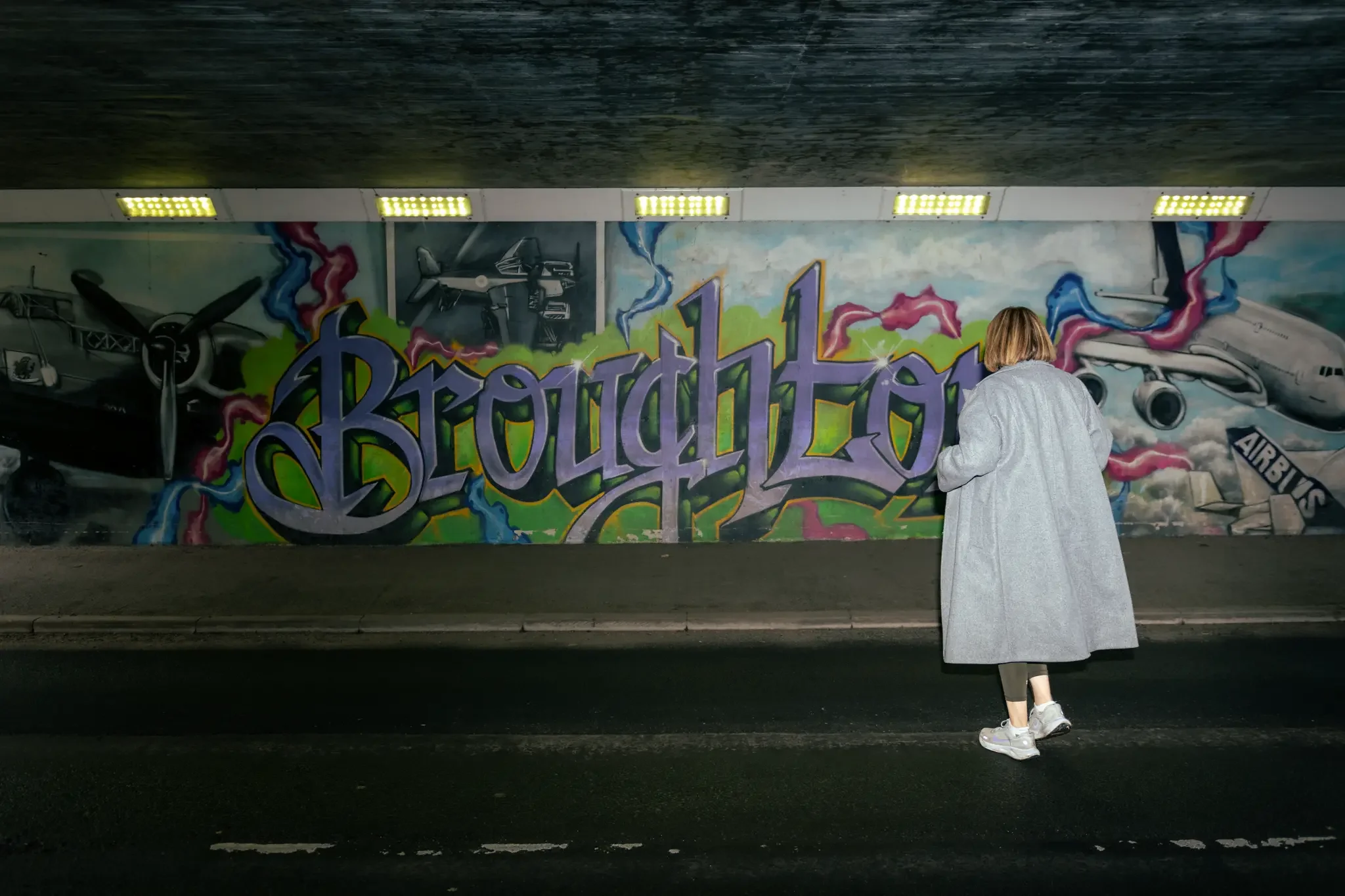Person in a gray coat standing in front of colorful graffiti mural under an overpass.