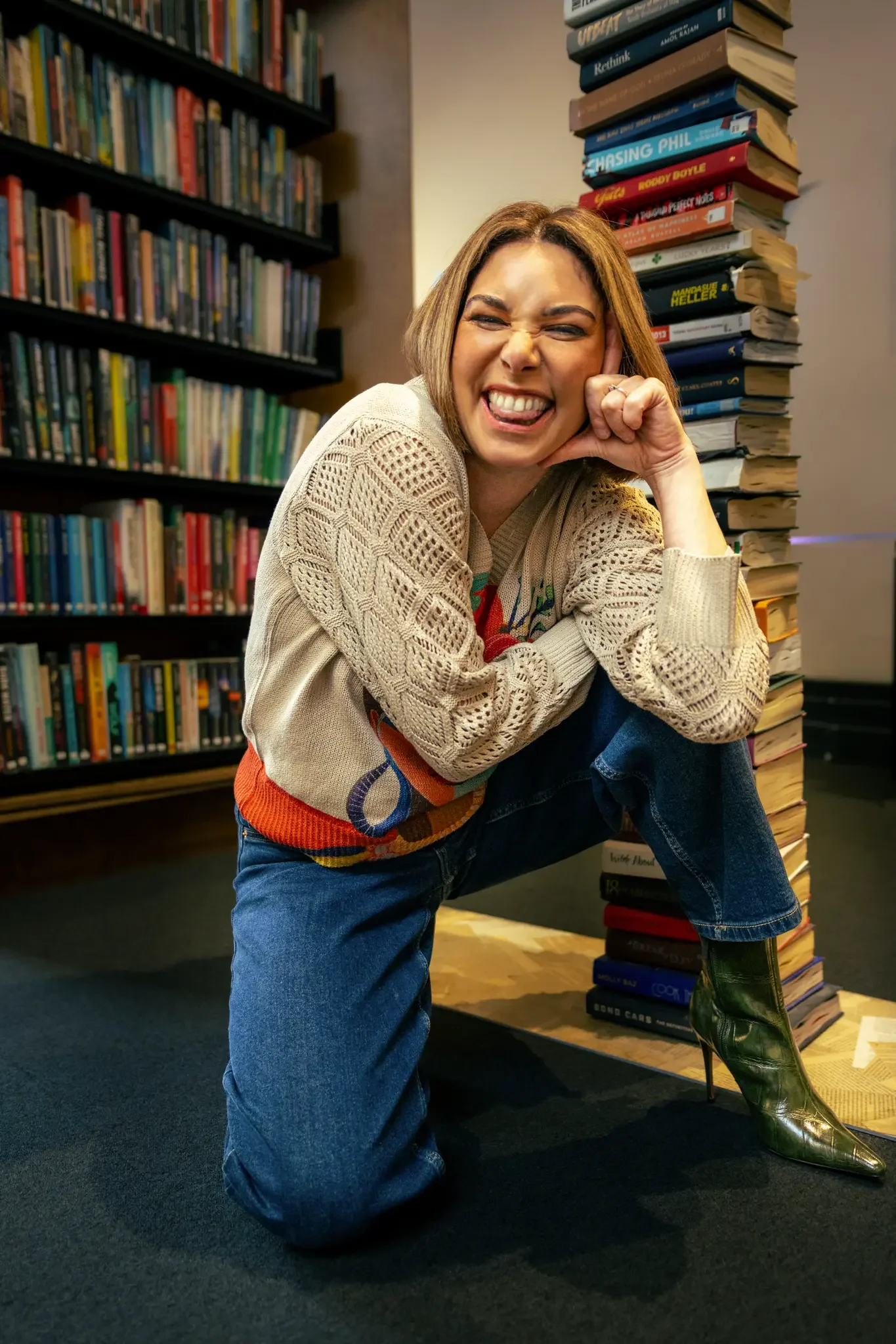A woman with light brown hair posing playfully in a bookstore, wearing a beige crochet sweater, blue jeans, and green high-heeled boots, surrounded by tall stacks of books and bookshelves filled with colorful books.