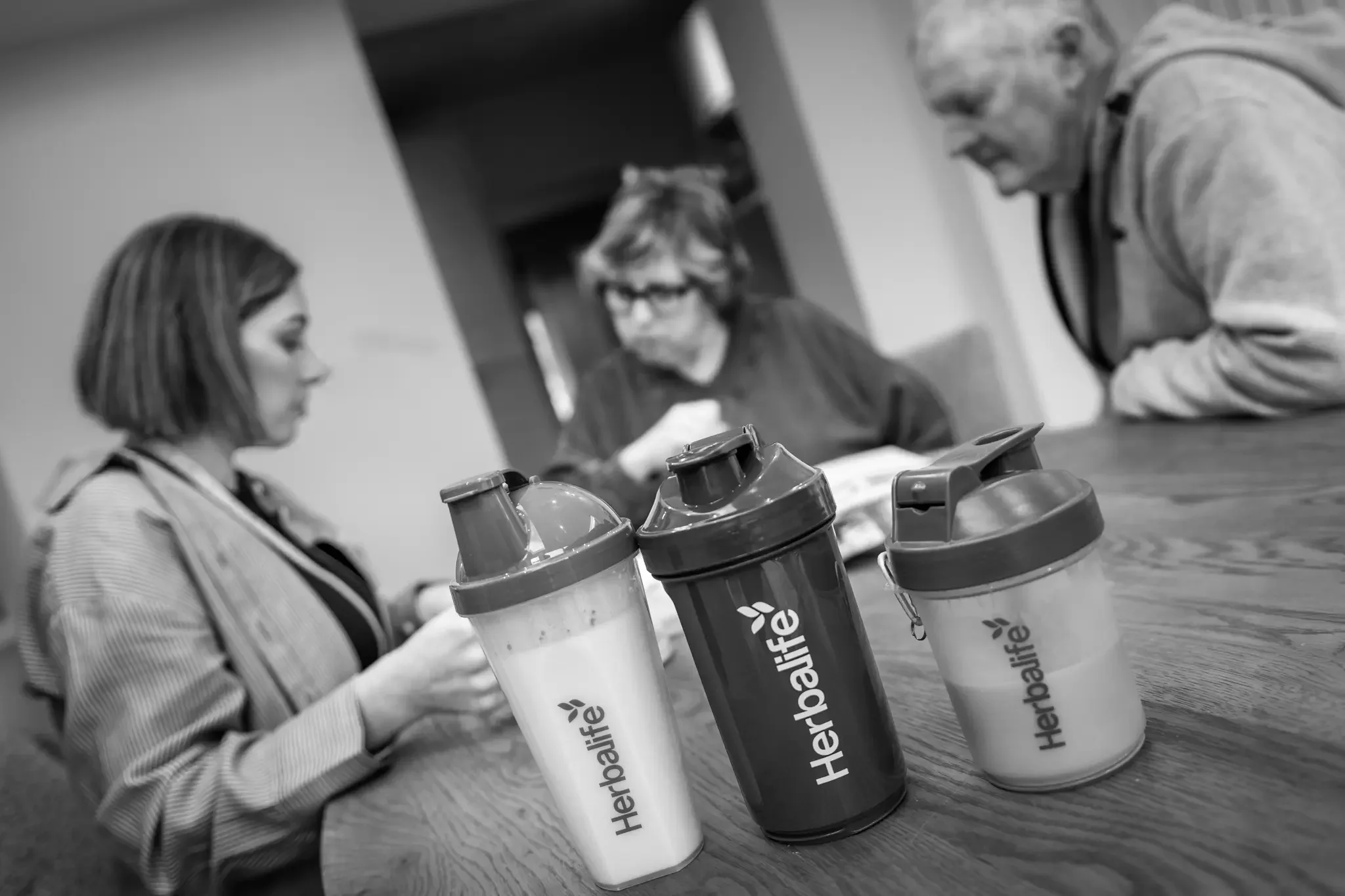 Three Herbafite protein shakers on a wooden table with three people in the background engaged in a discussion.