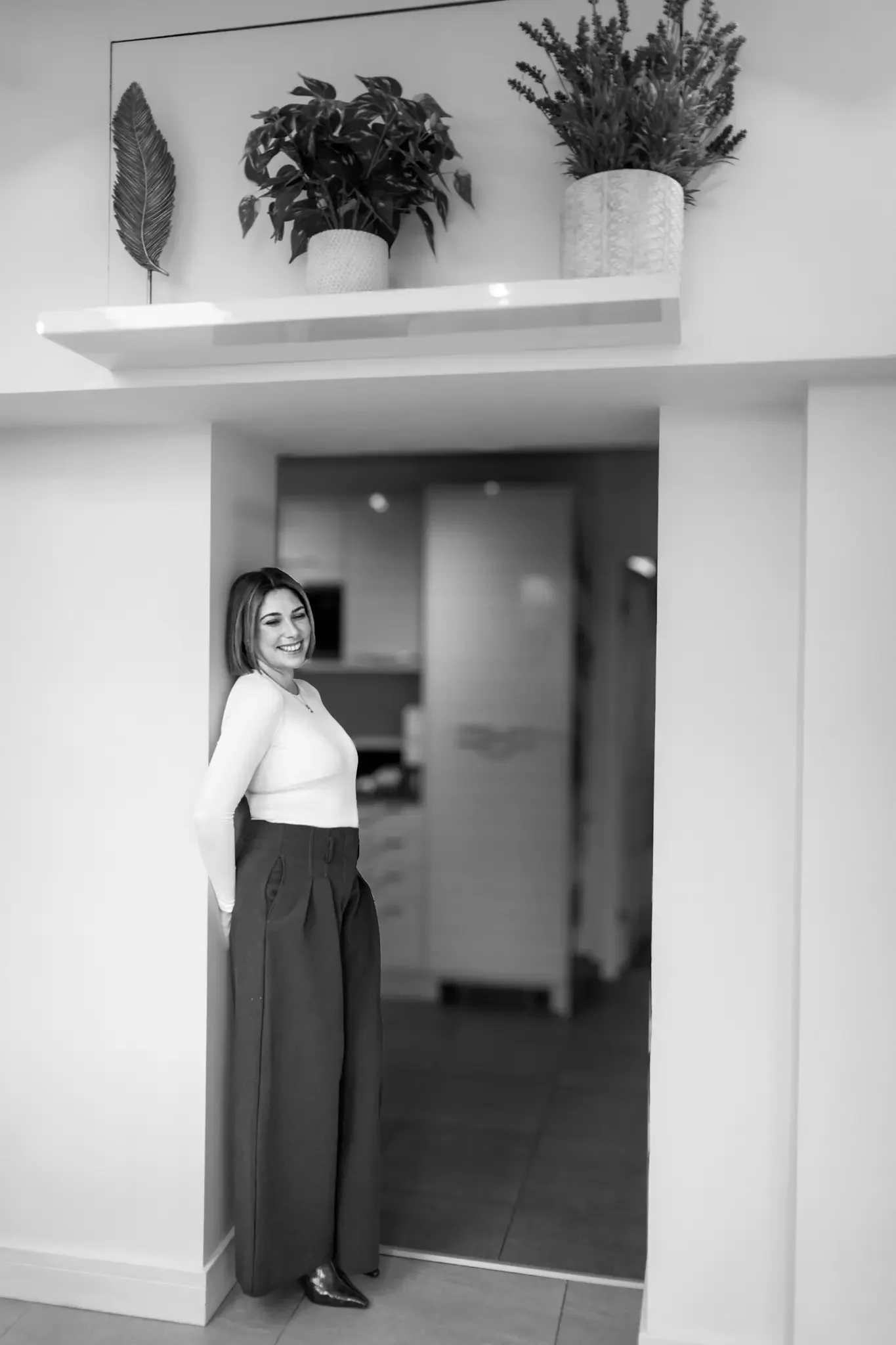 A woman smiling and posing in a doorway inside a house, with a wall shelf holding potted plants and decorative items above her.