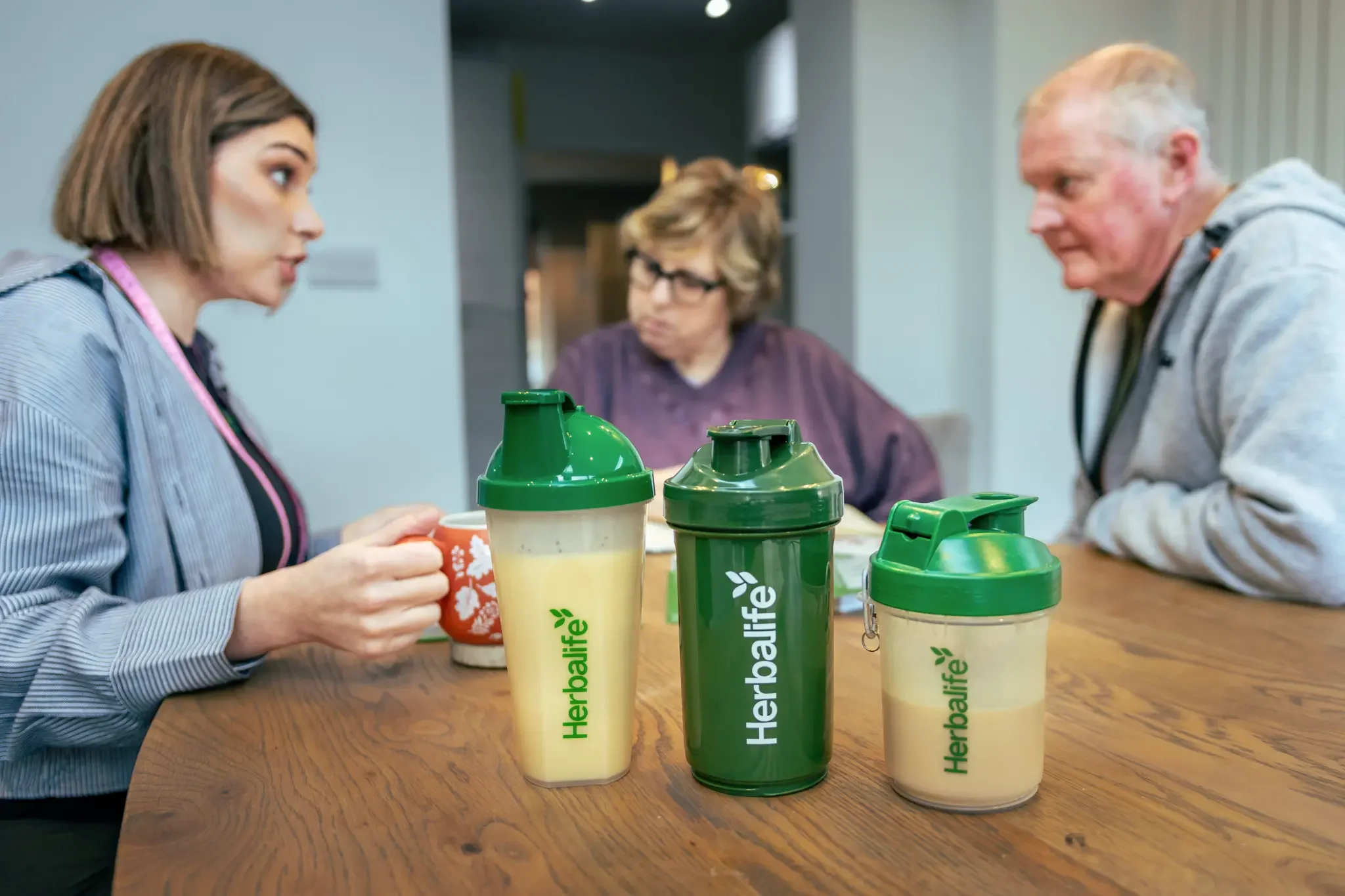 Three Herbalife protein shake bottles on a wooden table, with three people seated around, engaging in a conversation.