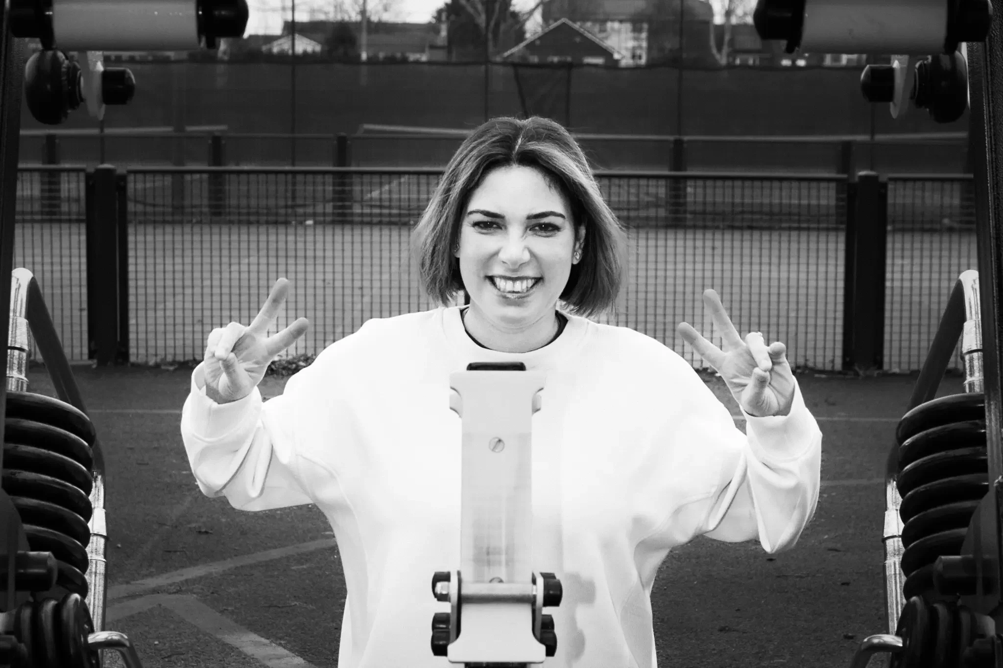 A young woman smiling and making peace signs with both hands at an outdoor sports court, with gym equipment and a fenced background.