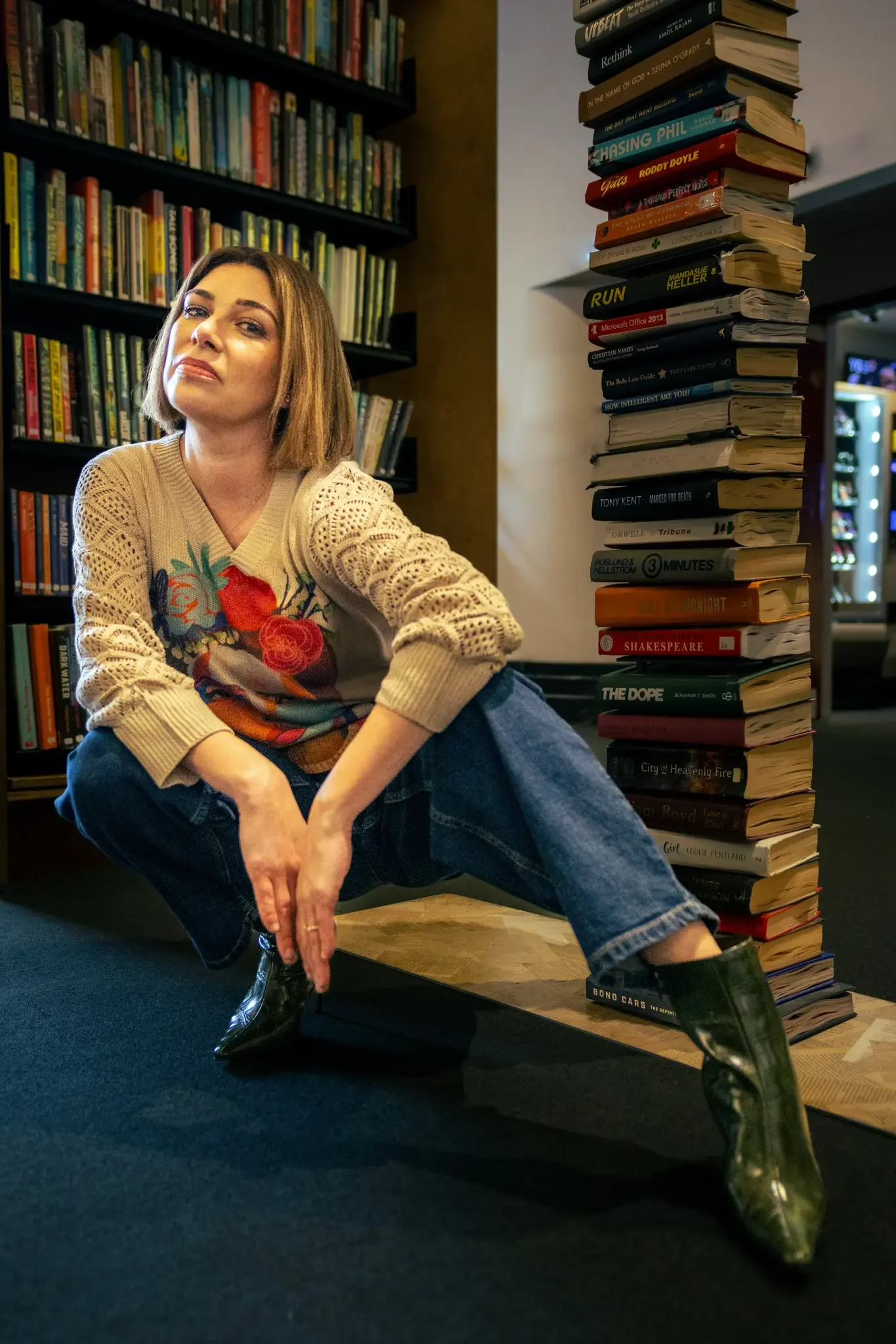 A woman with short brown hair sitting on the floor in a library, posing confidently with one leg extended, wearing a colorful sweater, blue jeans, and shiny black boots, with tall bookshelves and a stack of books in the background.