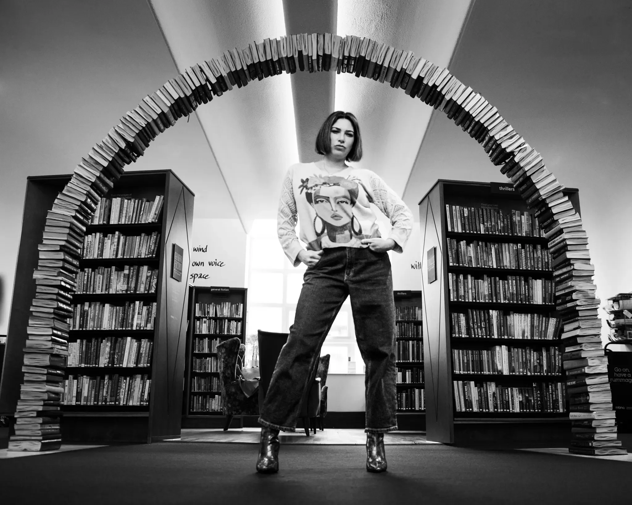 A woman standing in a bookstore, inside a large arch made of stacked books. She has shoulder-length hair, wears a graphic sweater, high-waisted jeans, and ankle boots. Bookshelves are visible in the background, along with a window letting in natural 