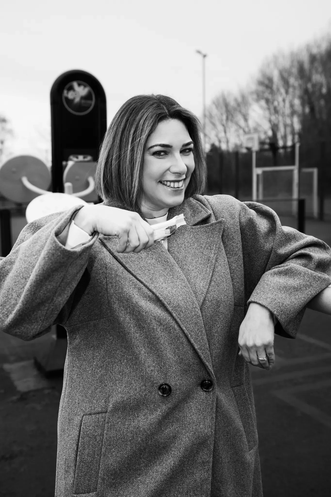 A woman in a gray coat smiling and holding a snack in her hand outdoors, with a playground in the background.