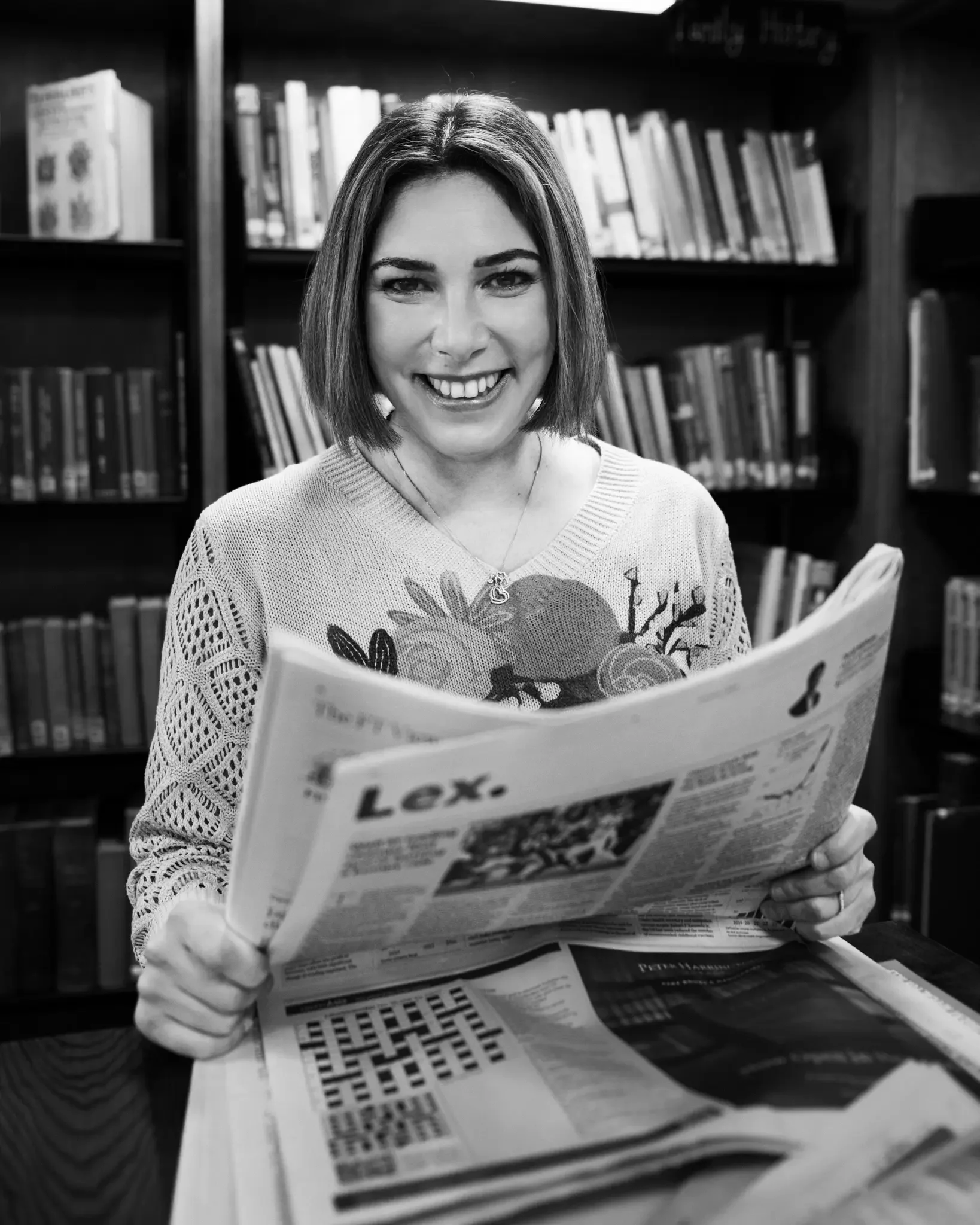 A woman with short hair smiling while reading a newspaper in a library. She is wearing a sweater with floral patterns and sitting at a table surrounded by bookshelves.
