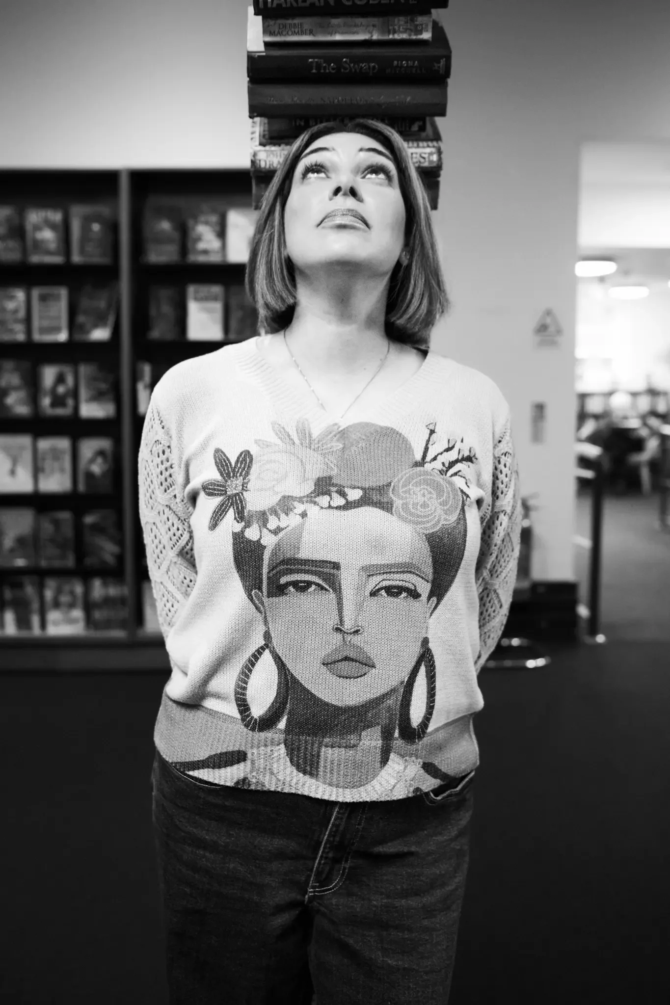A woman with short hair looks upward at a stack of books floating above her head in a bookstore or library.