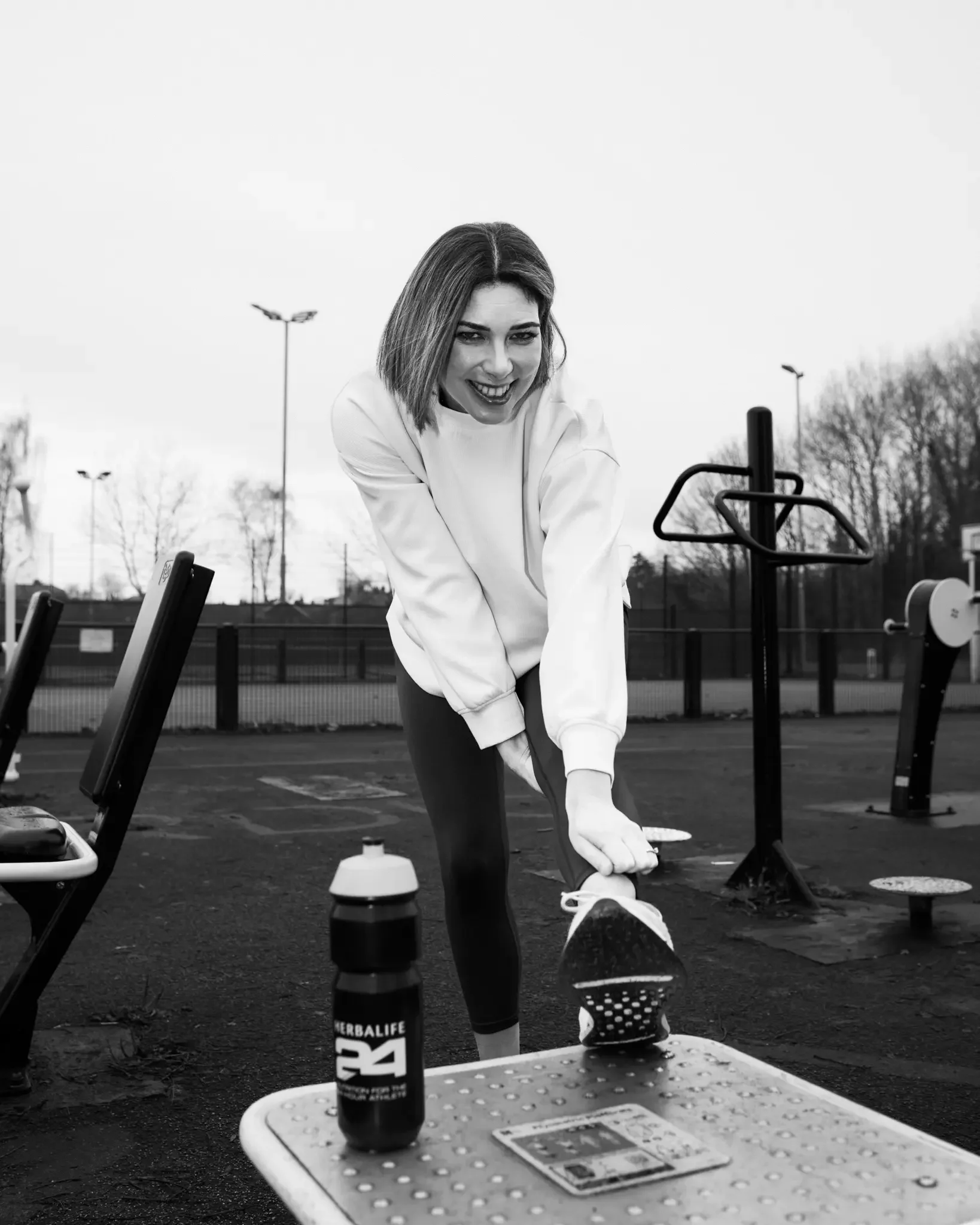 A woman with shoulder-length hair in athletic clothing, smiling as she stretches or exercises outside on a sports court with exercise equipment around her.