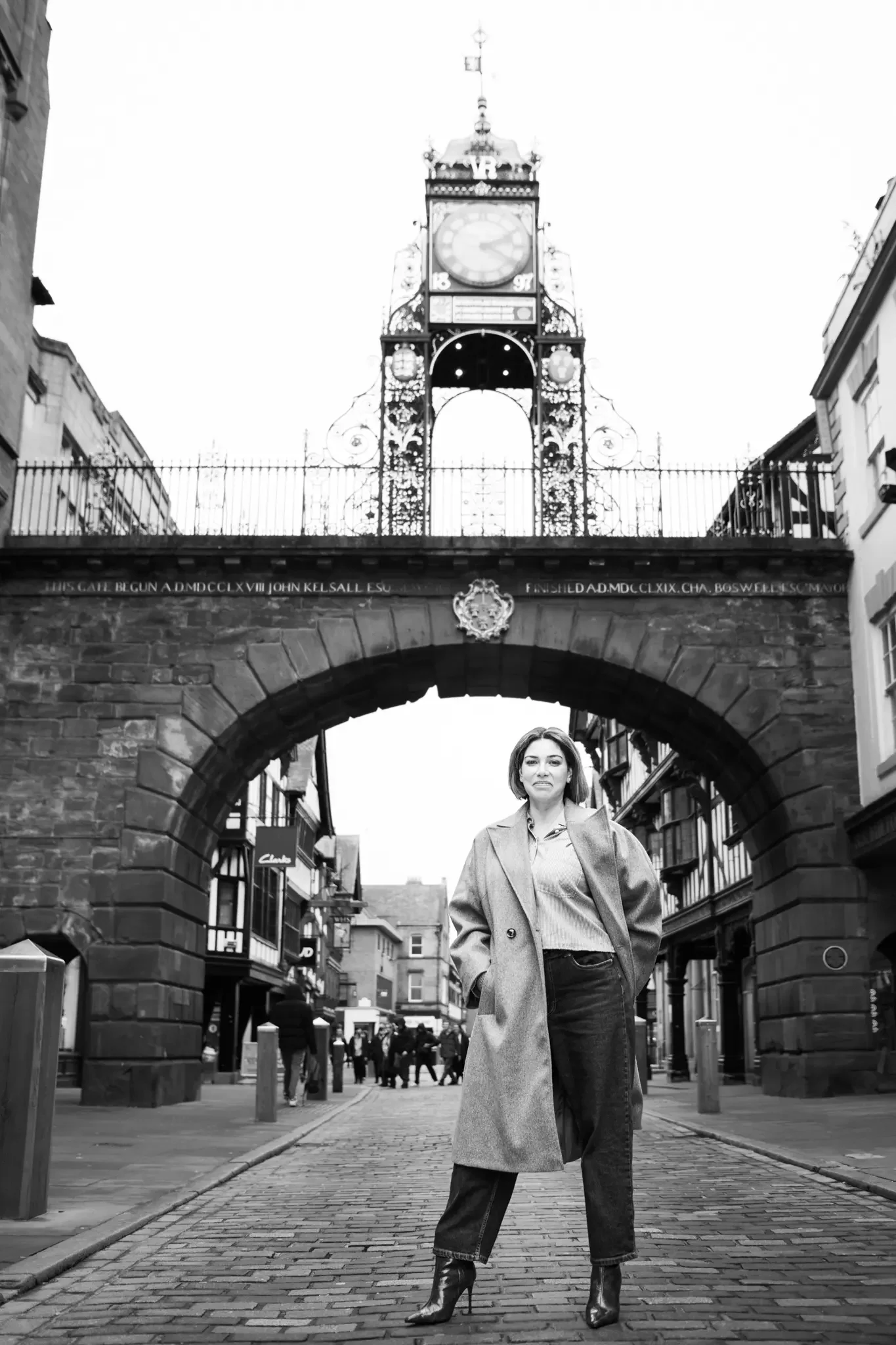 A woman standing beneath a historic gate with an ornate clock tower in the background on a cobblestone street.