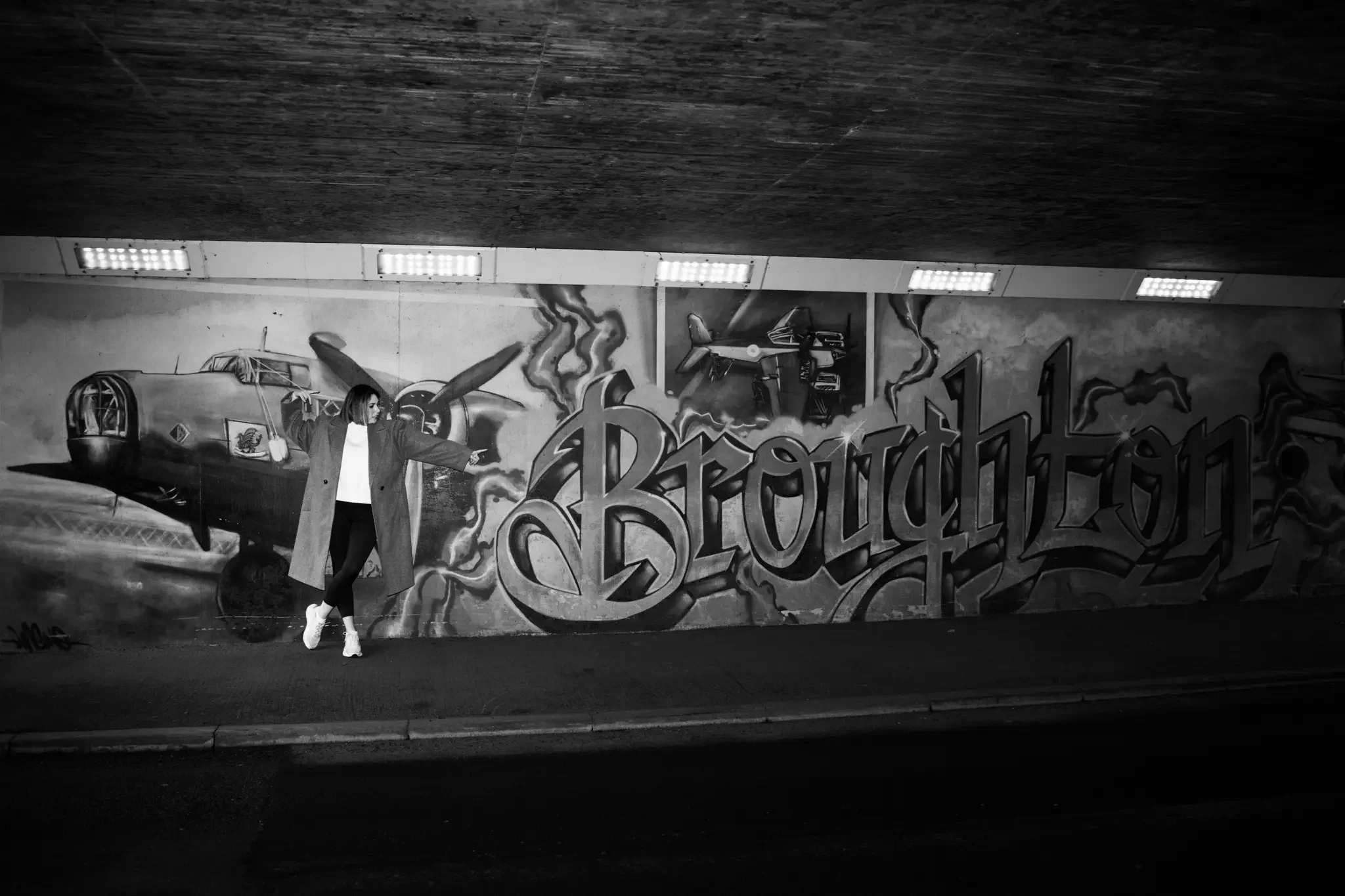 A woman in a coat and white boots standing in front of graffiti art in black and white. The graffiti features a vintage airplane and the word 'Broughston' in stylized letters.