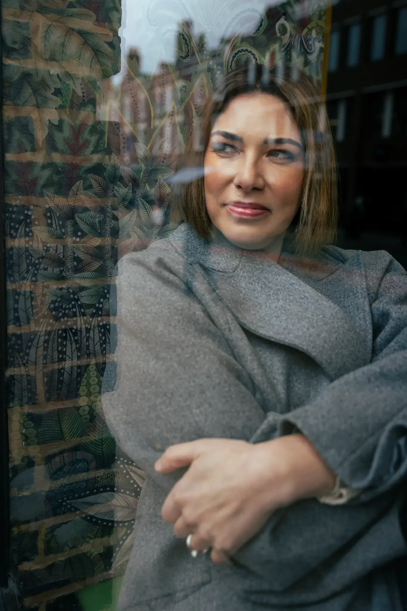 A woman with shoulder-length brown hair and a gray blazer is looking out a window with a thoughtful expression.