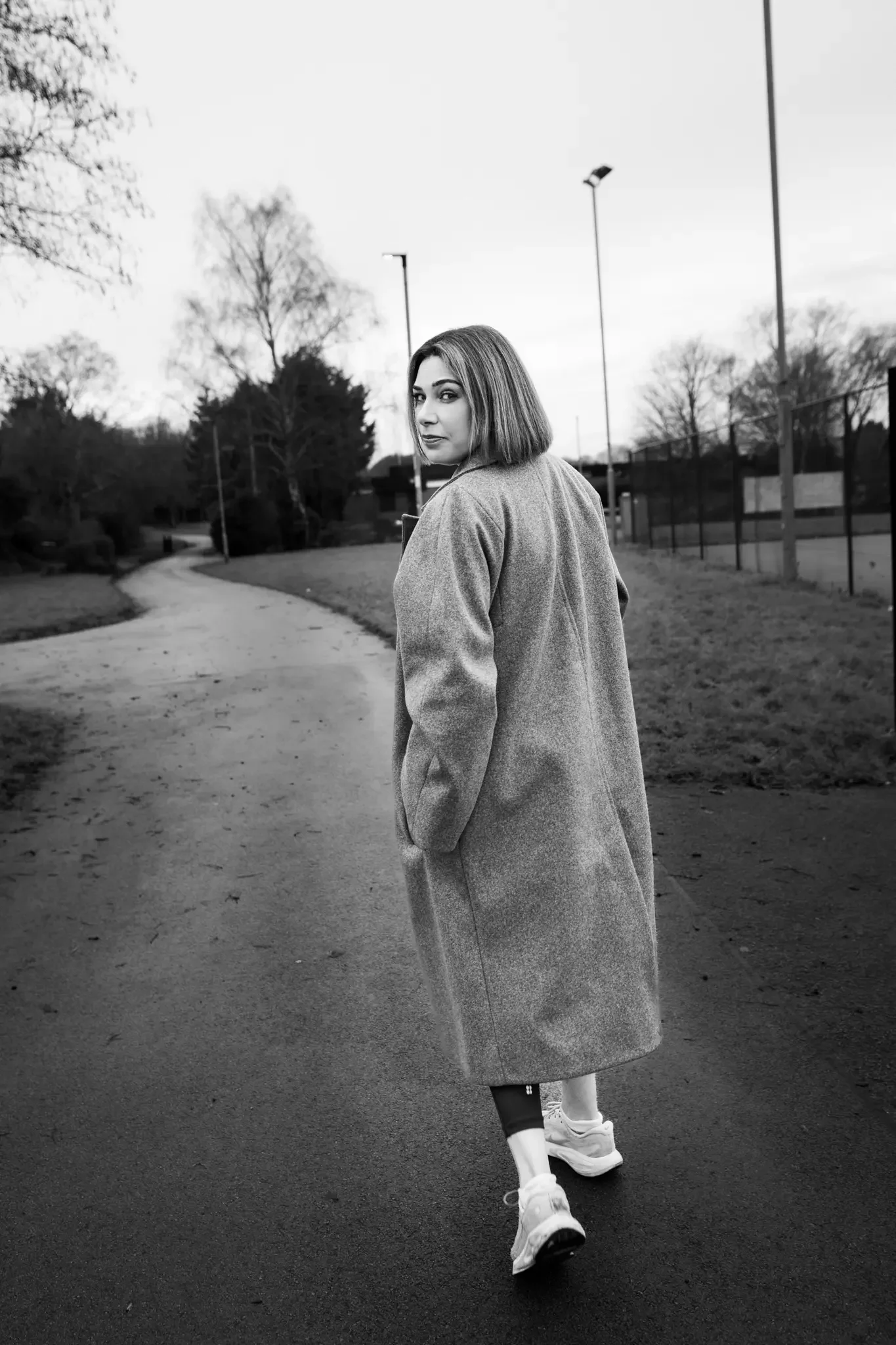 Black and white photo of a young woman with shoulder-length hair walking on a paved path in a park, wearing a long coat, sneakers, and leggings, looking over her shoulder.