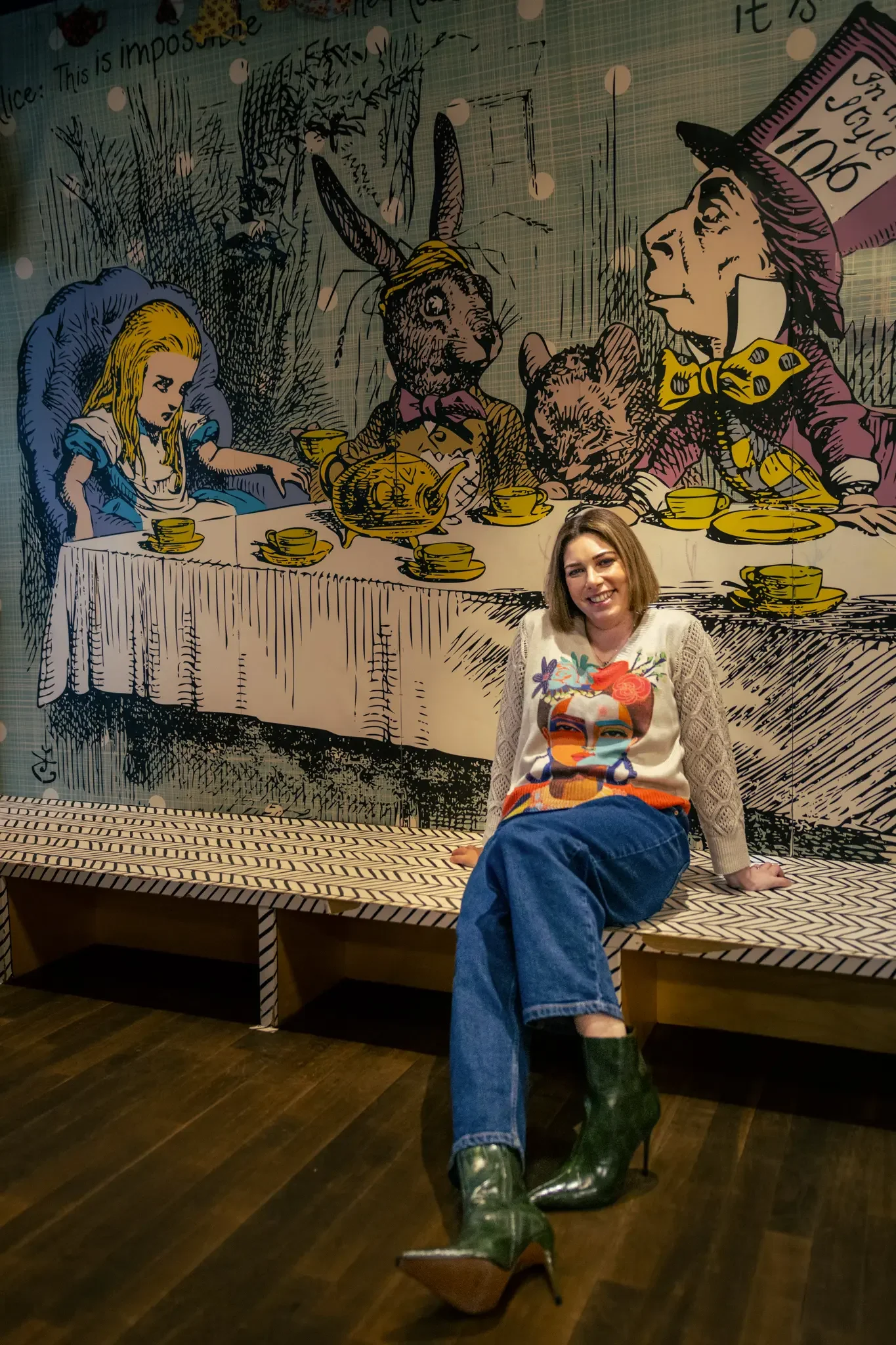 Woman sitting on a bench in front of a mural illustrating the Mad Hatter's Tea Party from Alice in Wonderland, featuring a girl, a rabbit, a cat, and the Mad Hatter.
