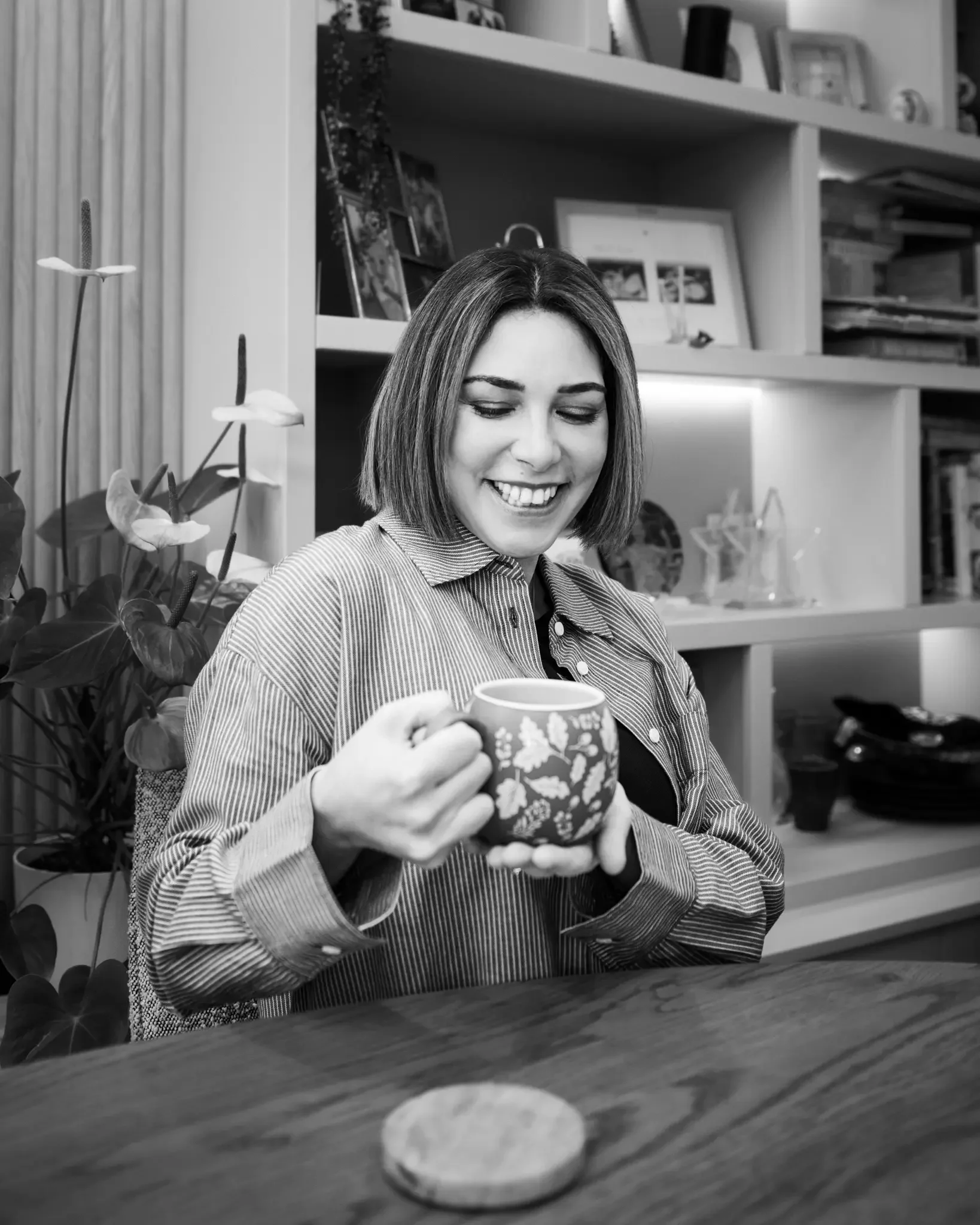 A woman with shoulder-length hair smiling while holding a patterned coffee mug in both hands, sitting at a wooden table. A small round coaster is on the table in front of her, and a bookshelf with framed photos and decorative items is in the backgrou
