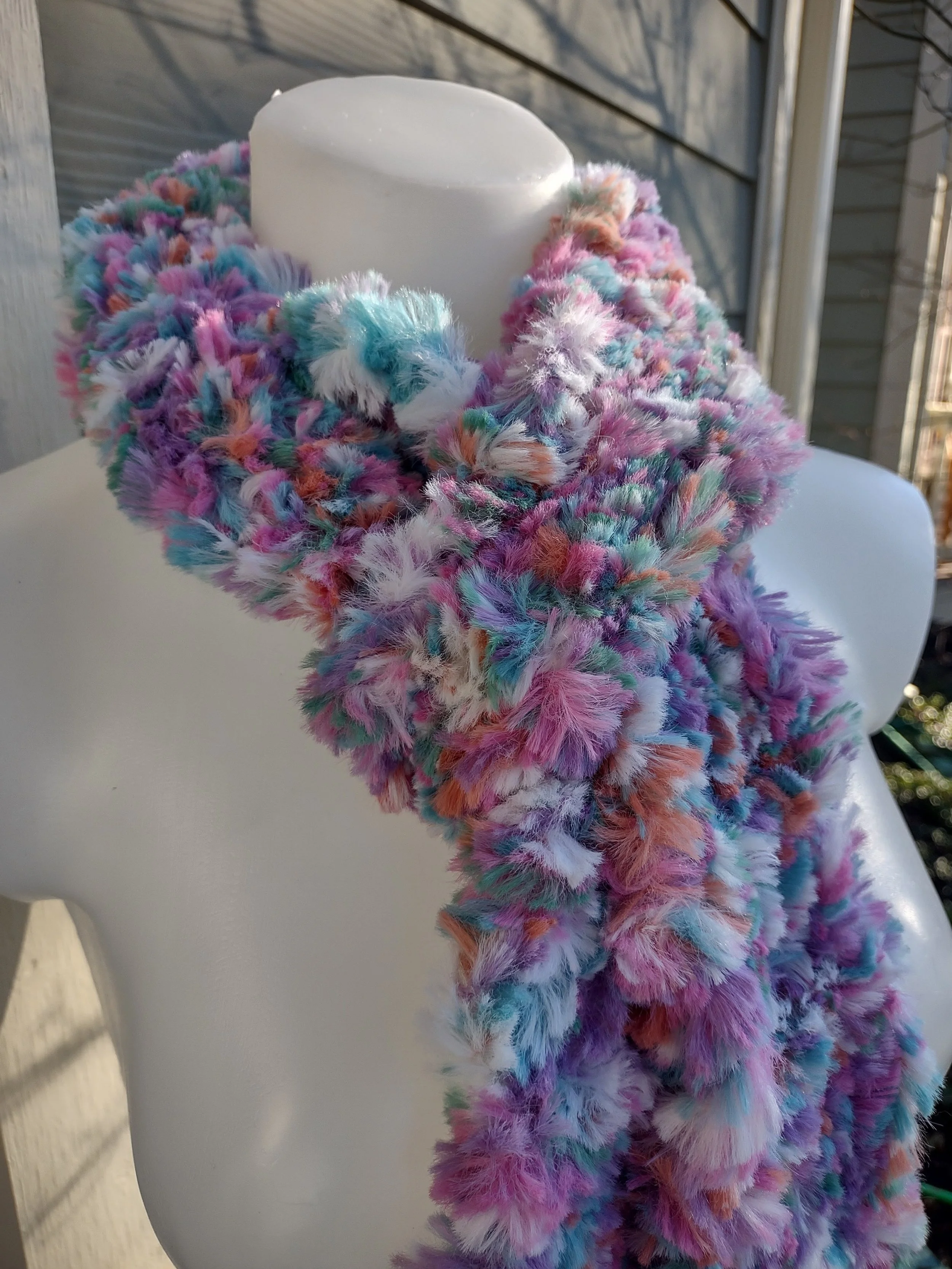 A close-up view of a pastel knit scarf wrapped around a mannequin’s neck, highlighting the fluffy texture and blend of pink, teal, purple, white, and orange fibers.