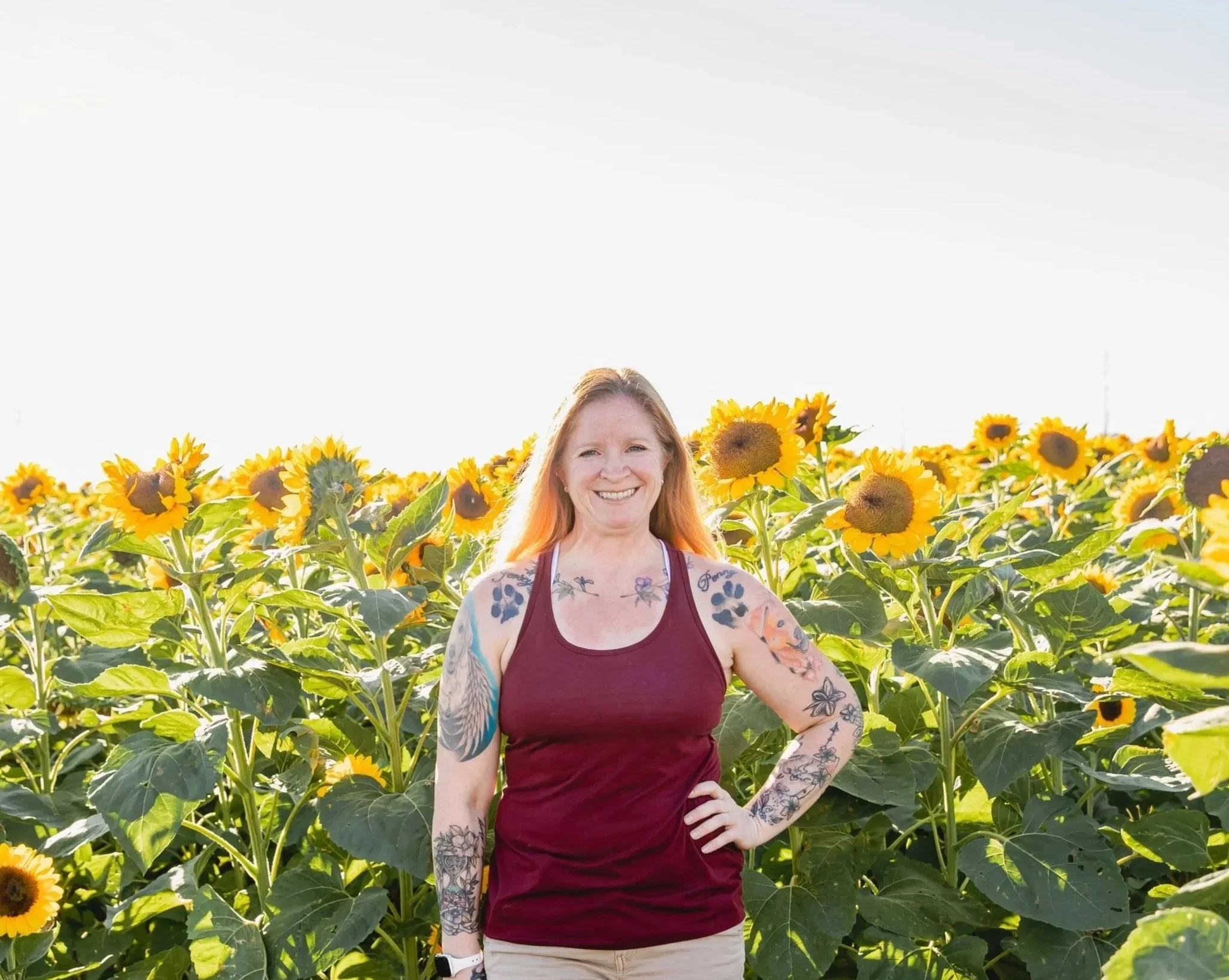 A female dog trainer and holistic pet health coach with dog tattoos standing in a sunflower field during daytime.