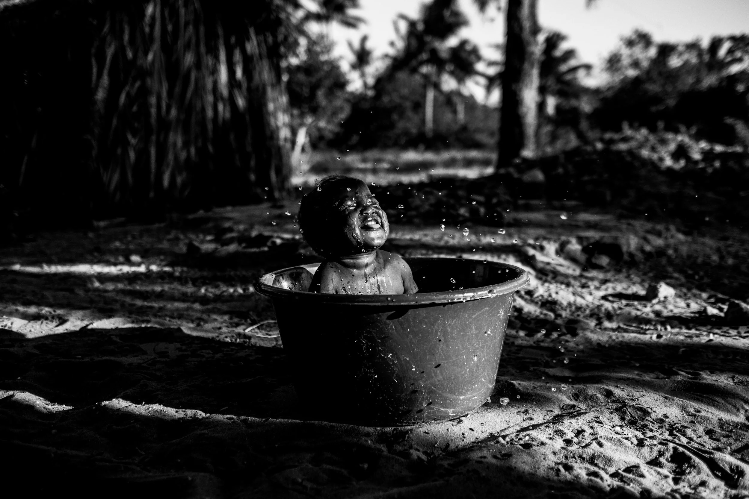 Therapy in Kenya.  Child playing in a water bucket outdoors, splashing water with a joyful expression, surrounded by natural trees and sandy ground, in black and white.