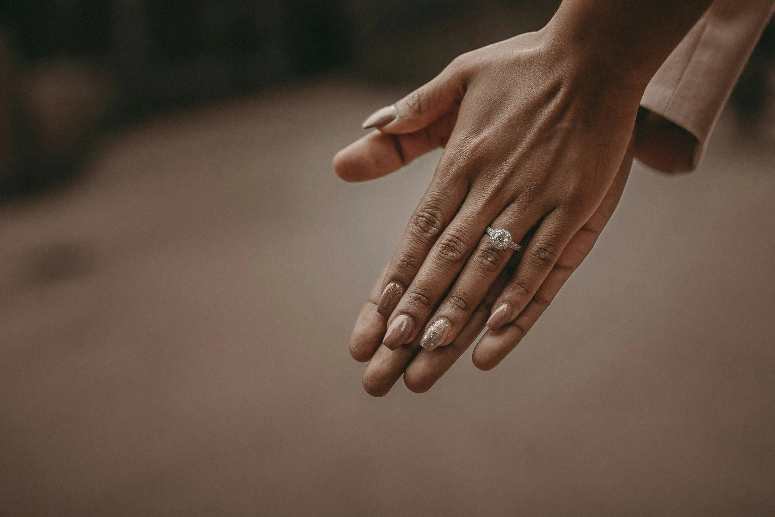 Therapy in Kenya.  A close-up of a woman's hand wearing a diamond engagement ring, with manicured nails painted in nude and glitter designs, against a blurred background.